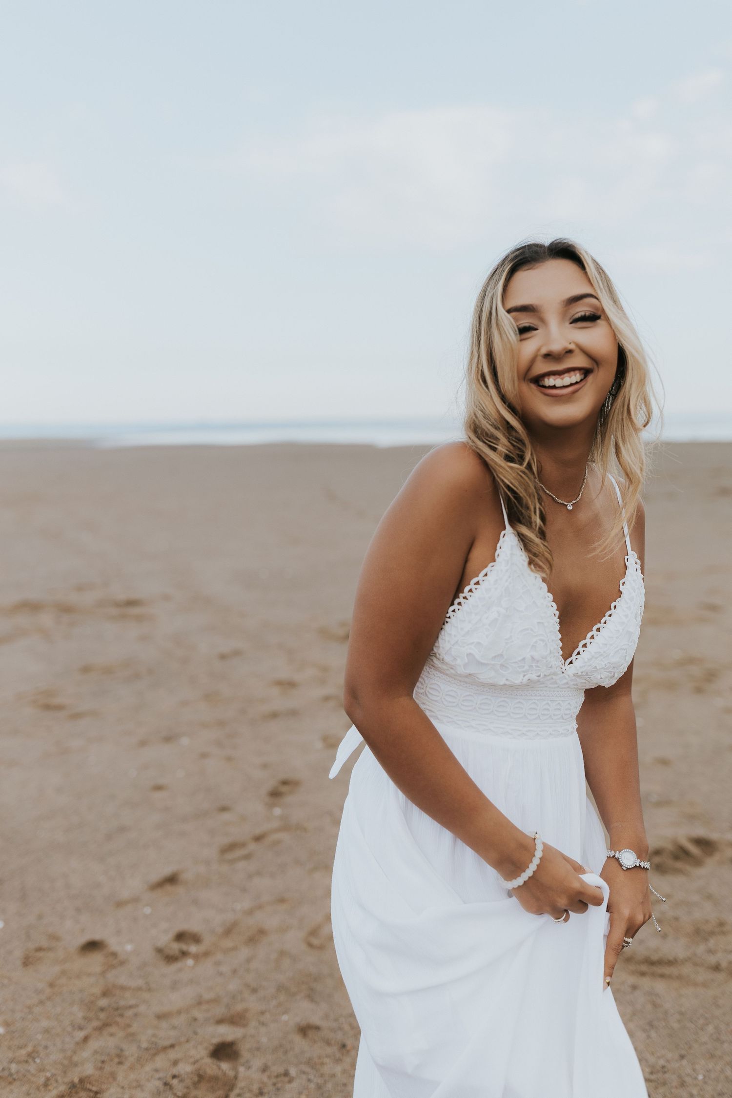 Laughing young woman in white lace sundress stands on sandy beach holding her dress in the breeze.