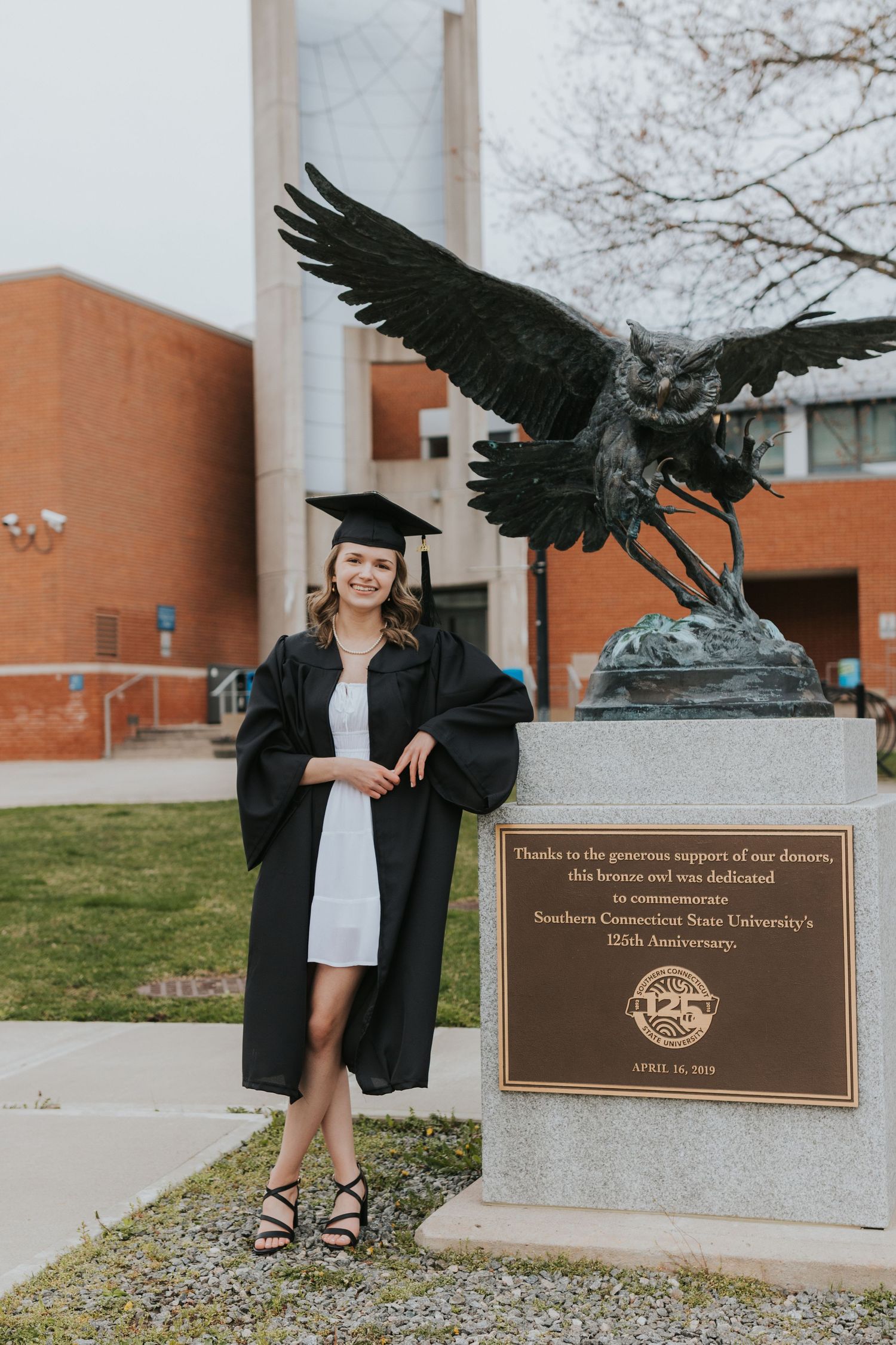 Female graduate in cap and gown poses beside eagle statue at Southern Connecticut State University.