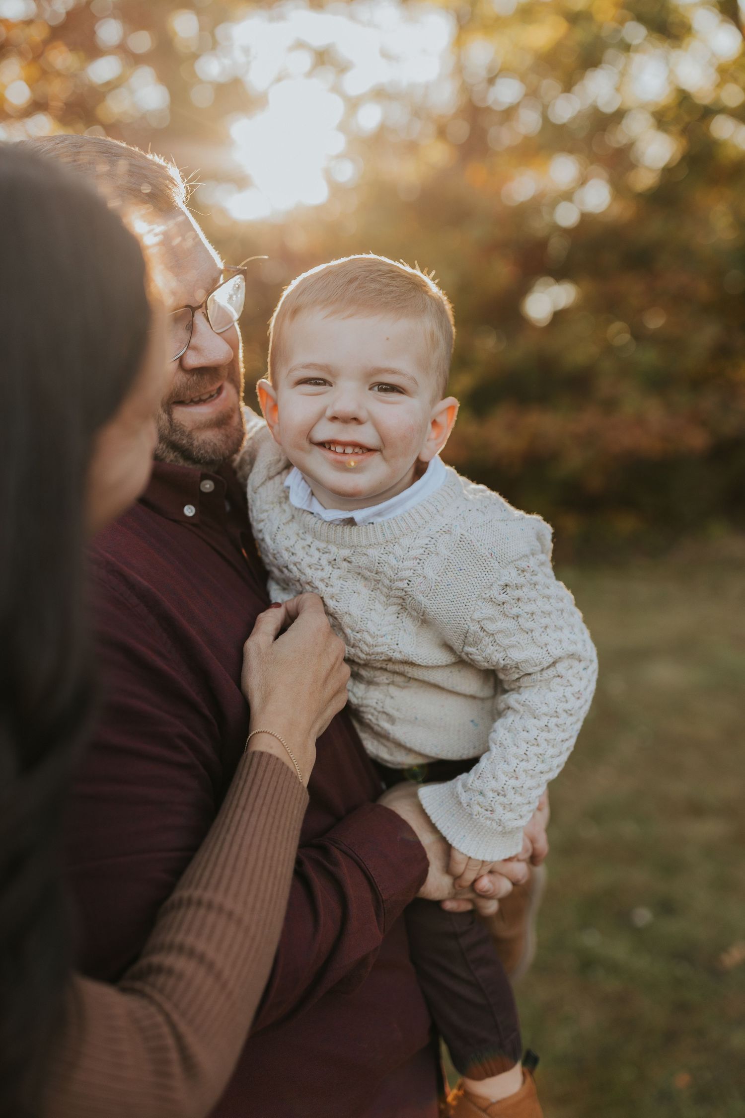 Smiling baby boy in cream sweater held by parents during warm autumn sunset family portrait session.