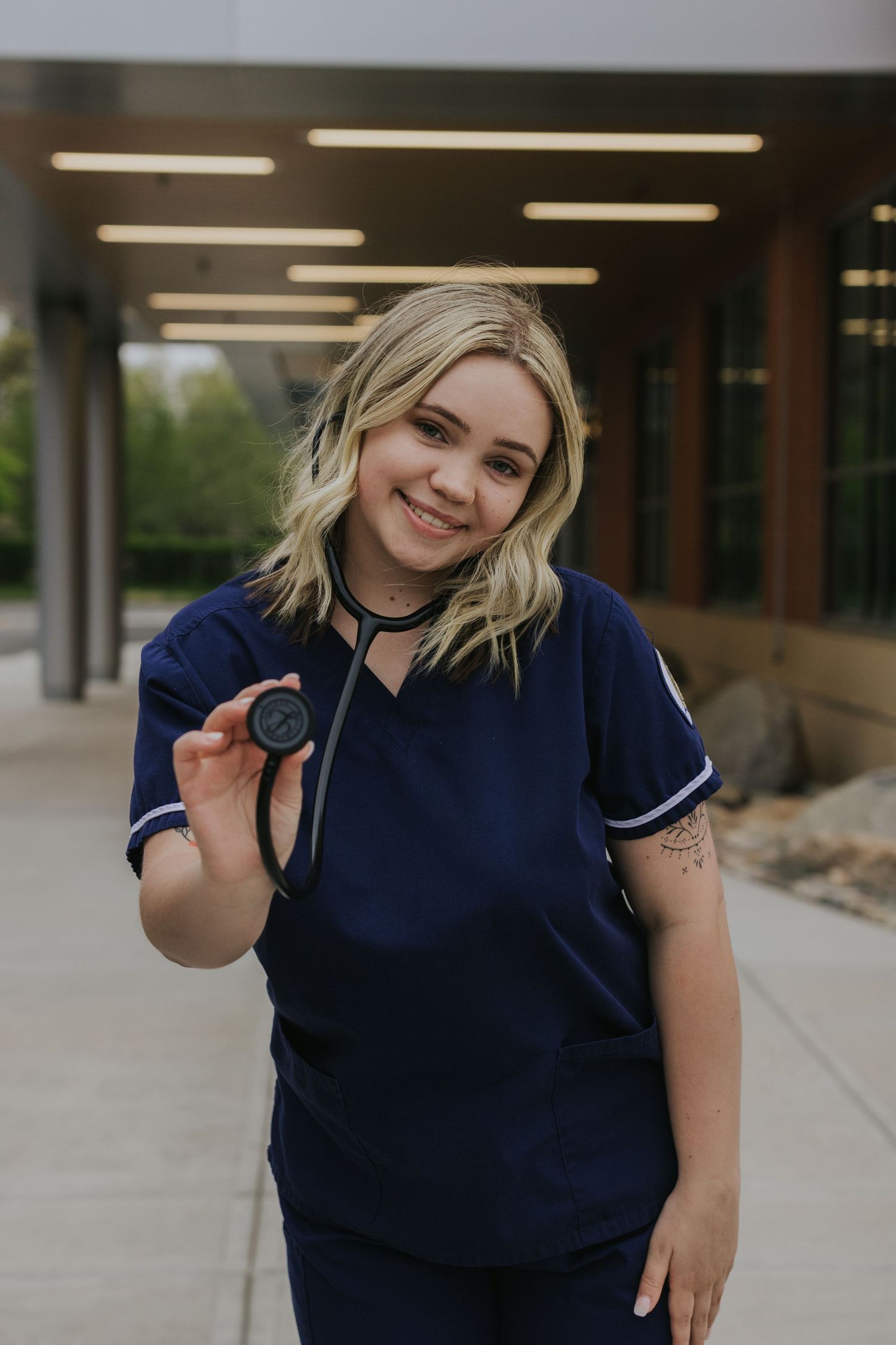Nursing student in navy scrubs holding stethoscope toward camera outdoors.