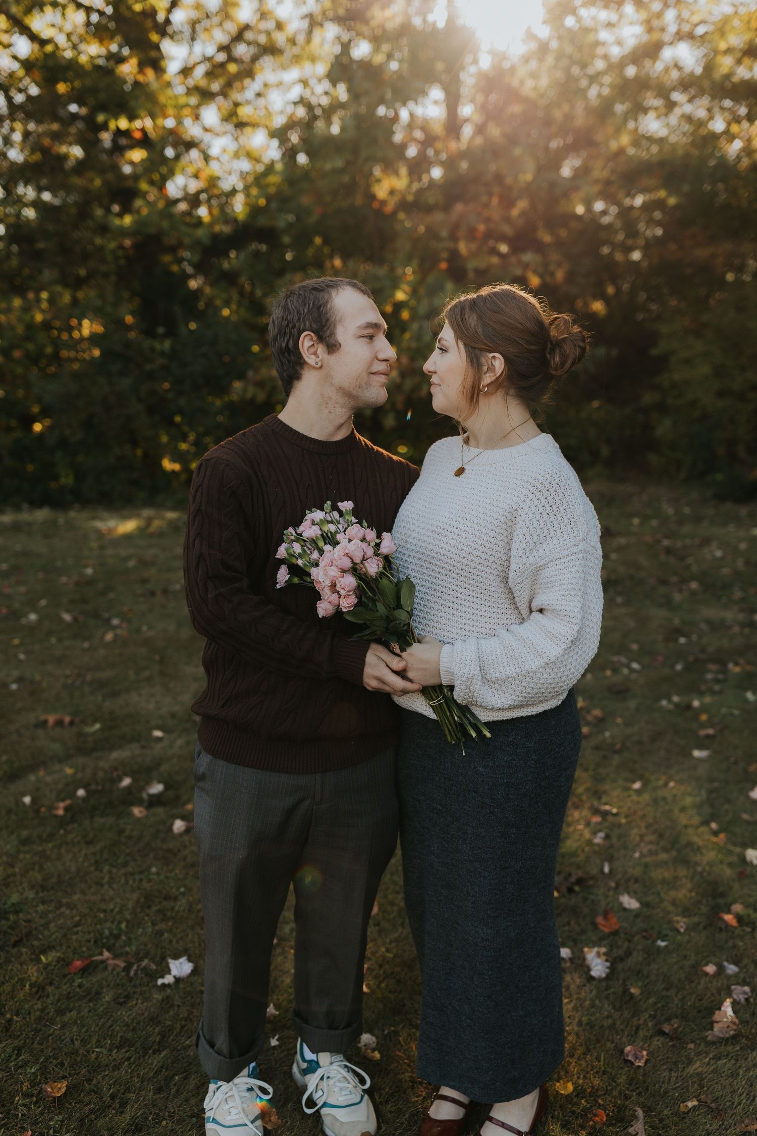 Couple sharing romantic nose-to-nose moment holding pink bouquet during golden hour outdoor maternity session.
