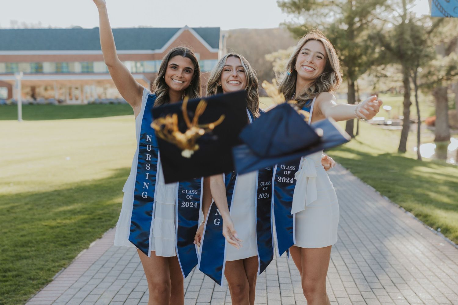 Three graduating women celebrating outdoors in caps and gowns with blue sashes.