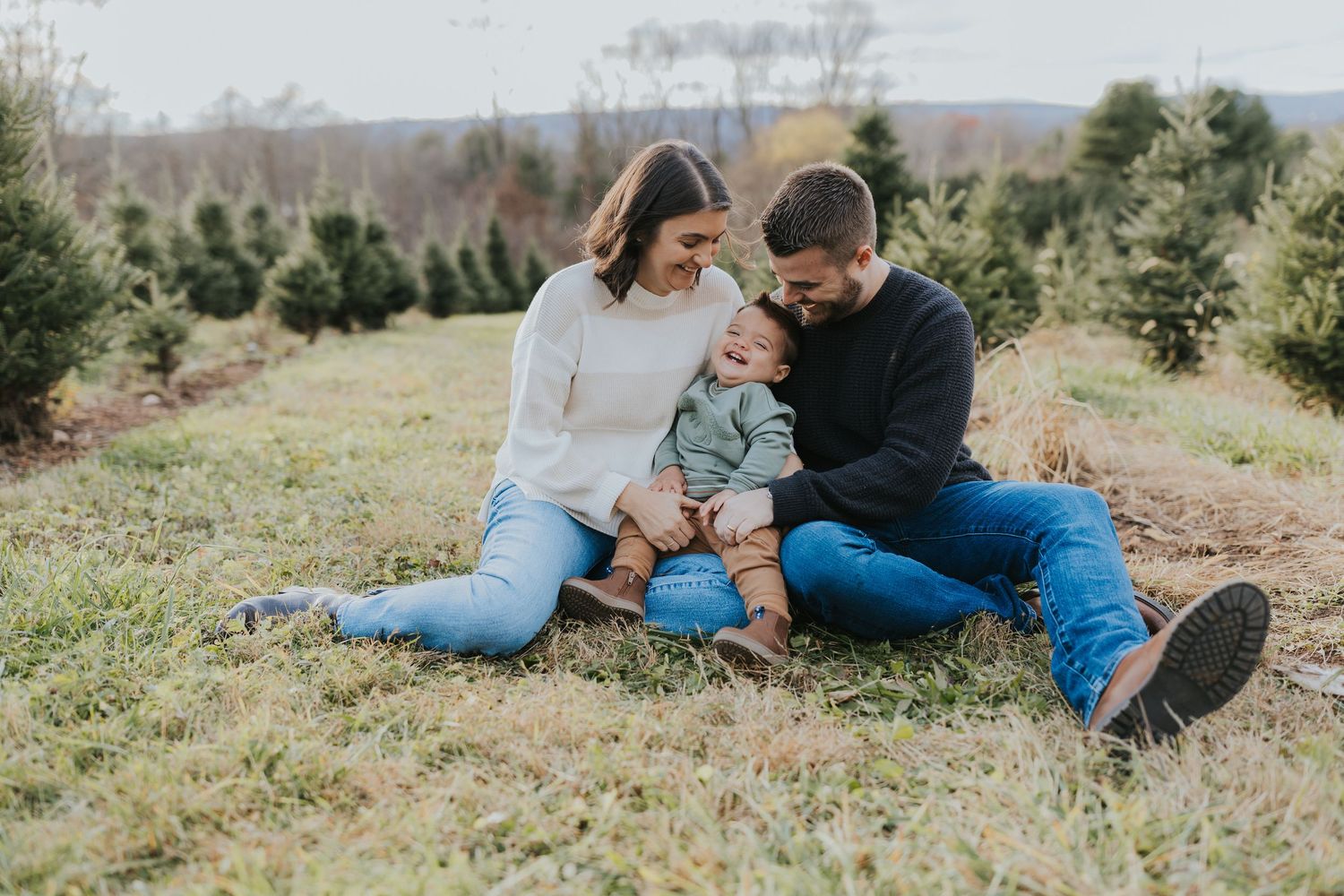 Young family sits together on grass at a Christmas tree farm, parents smiling at their baby between them.