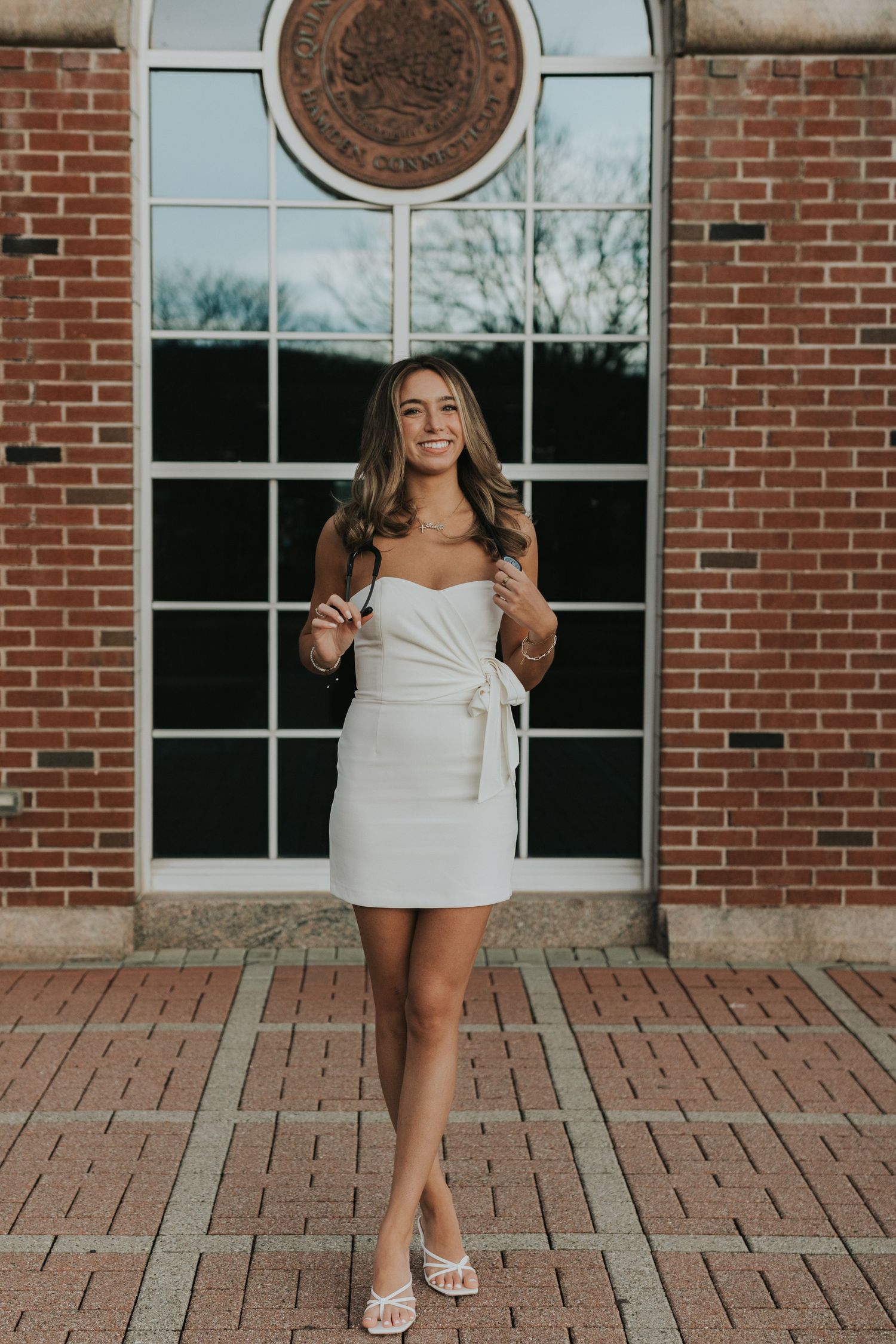 Young woman in white strapless mini dress posing in front of brick building with large window.