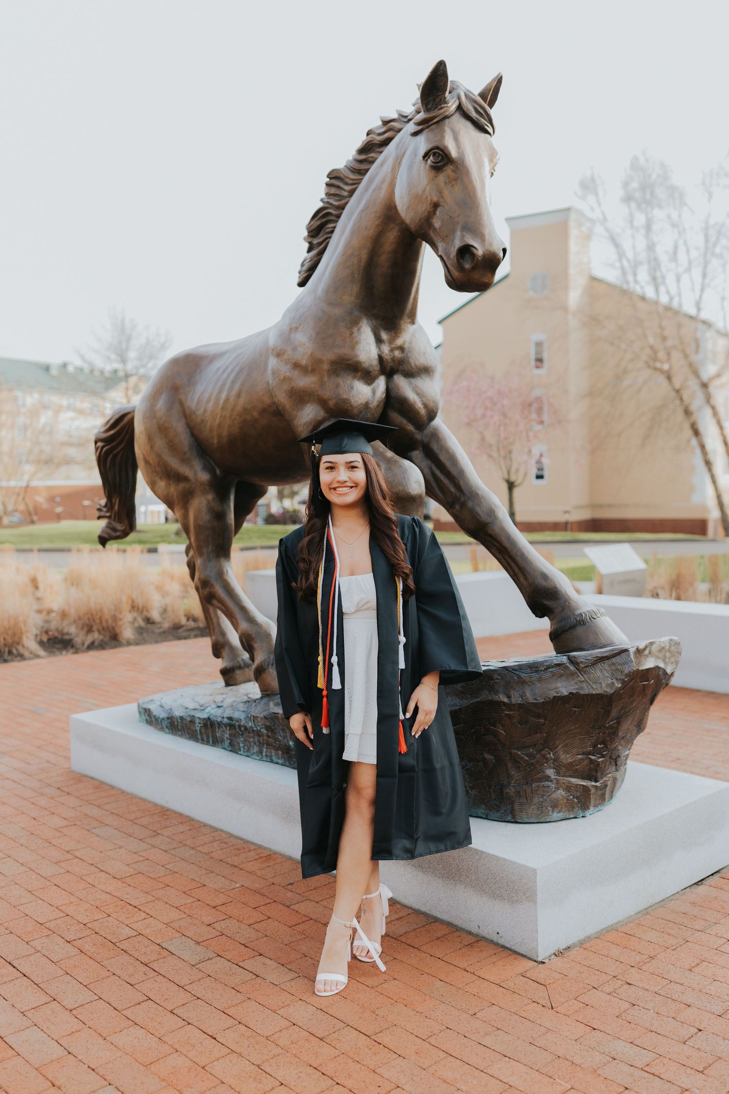 Graduate in cap and gown posing in front of bronze horse statue on college campus.
