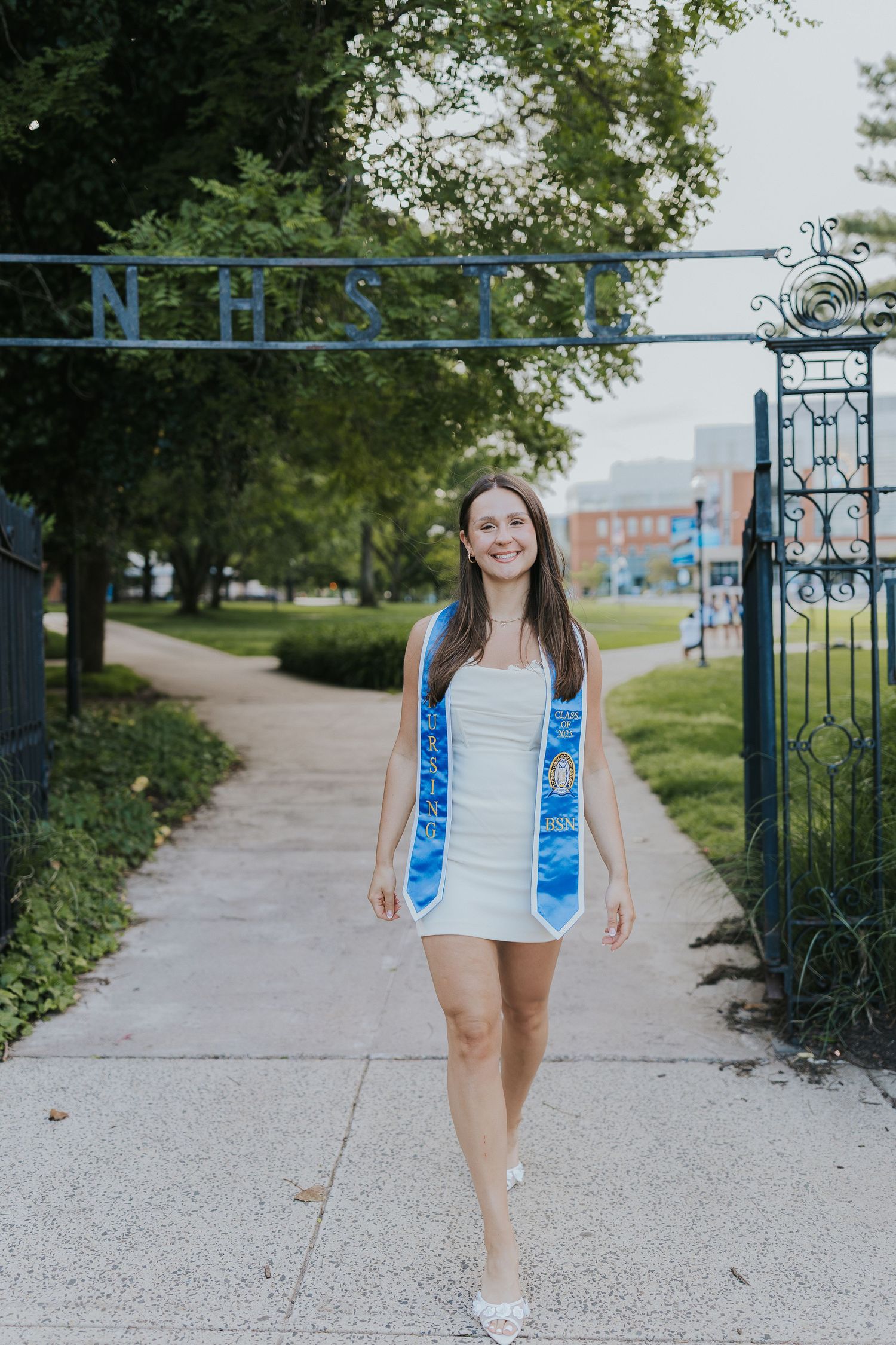 Nursing graduate walks through NHST campus gate wearing blue graduation sash.