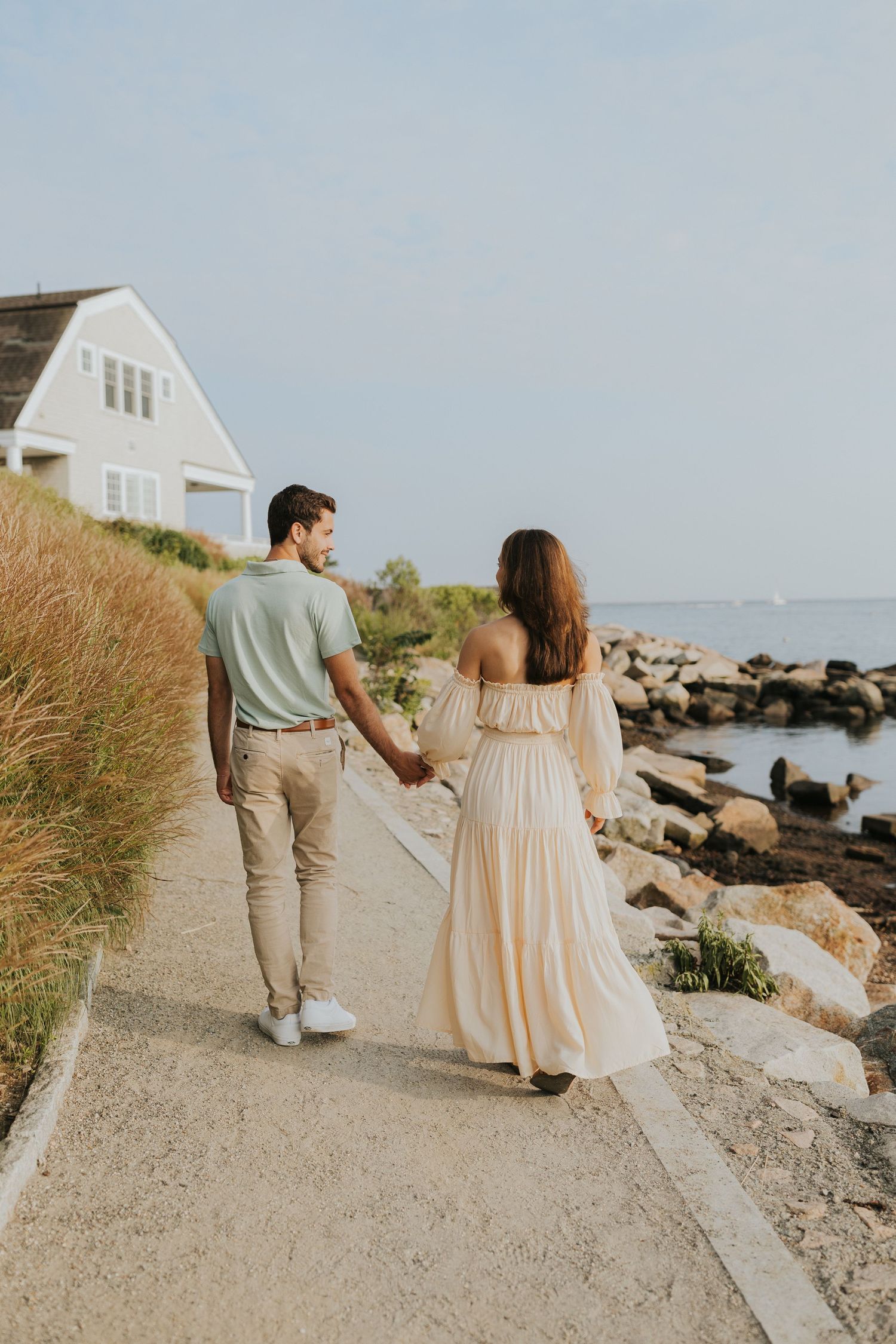 Couple walking hand in hand along a coastal path toward the ocean, viewed from behind at golden hour.