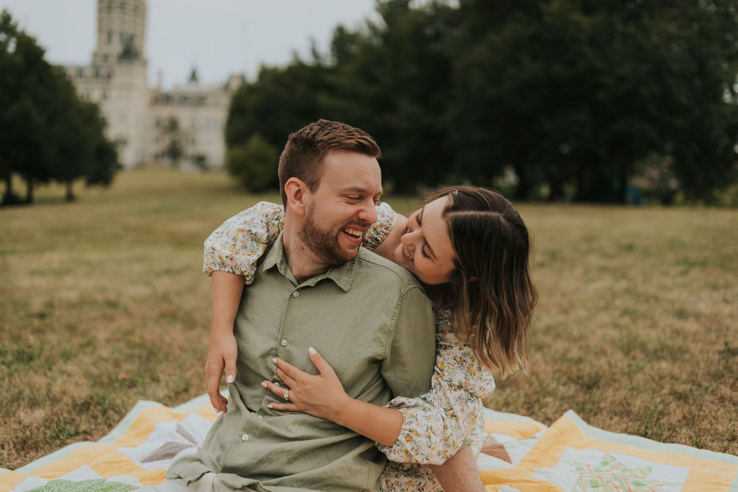 Joyful couple laughing and embracing on a picnic blanket in a grassy park setting.