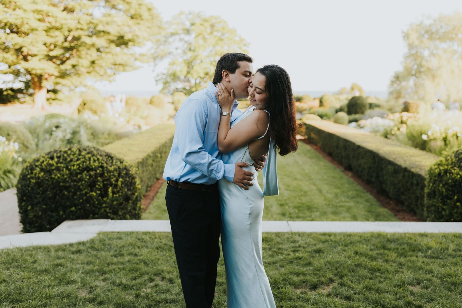 Couple sharing a romantic kiss in a lush garden setting, man in blue shirt embracing woman in light green dress.