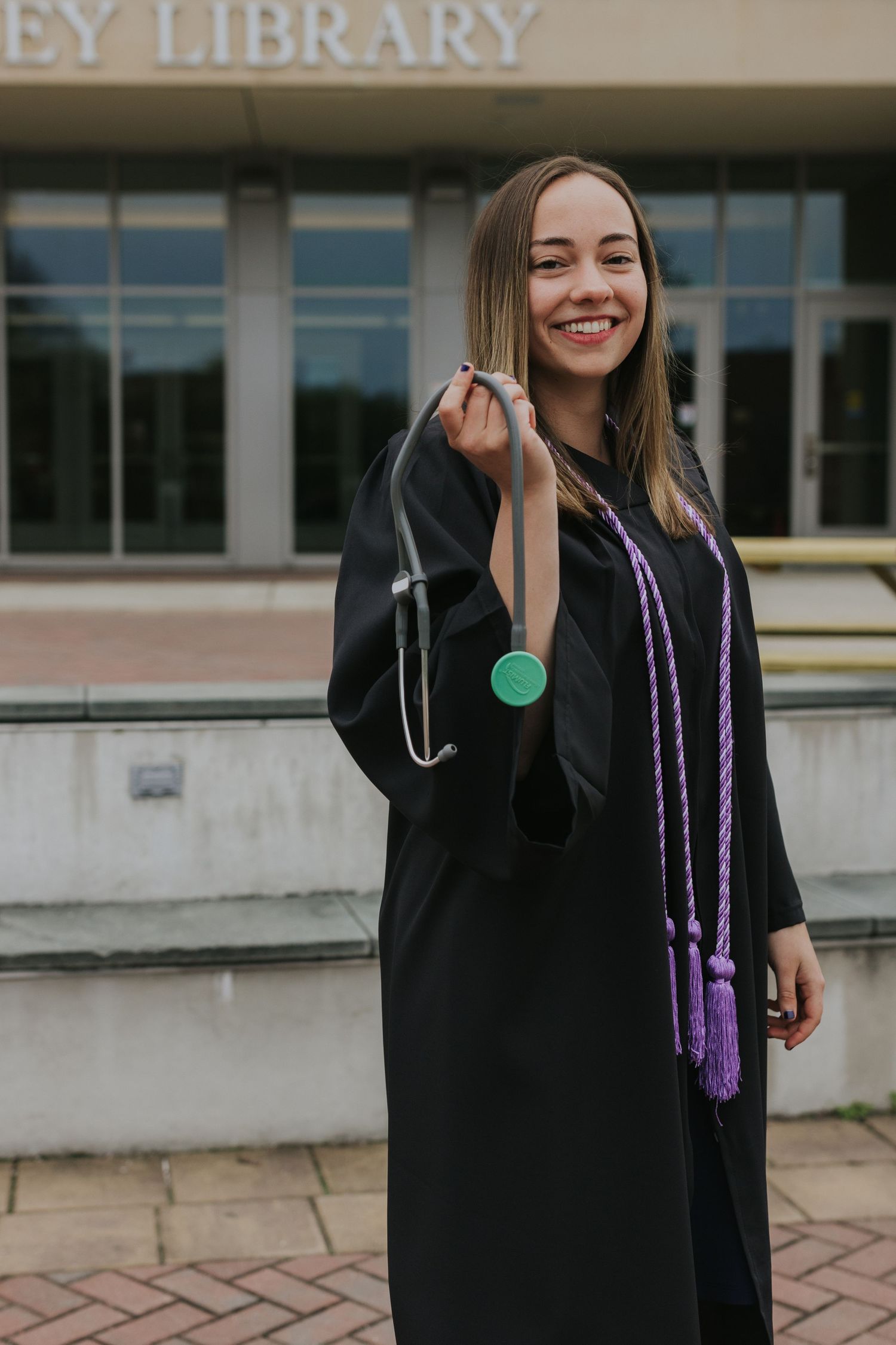 Smiling female nursing graduate in black gown holds stethoscope toward camera outside Buley Library building.