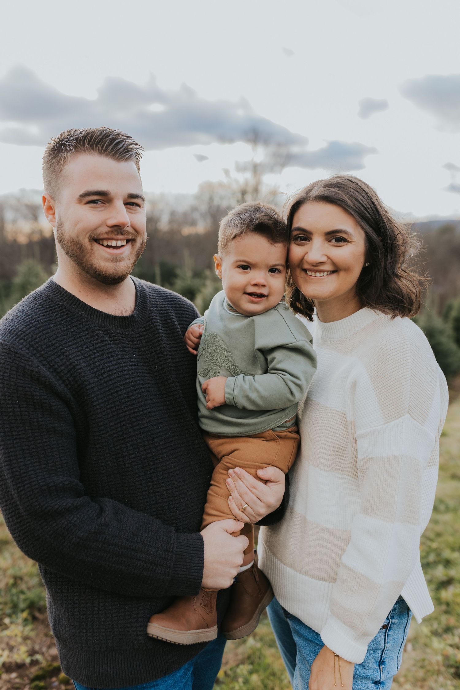 A happy young family poses together at a Christmas tree farm, dad holding their infant son outdoors.