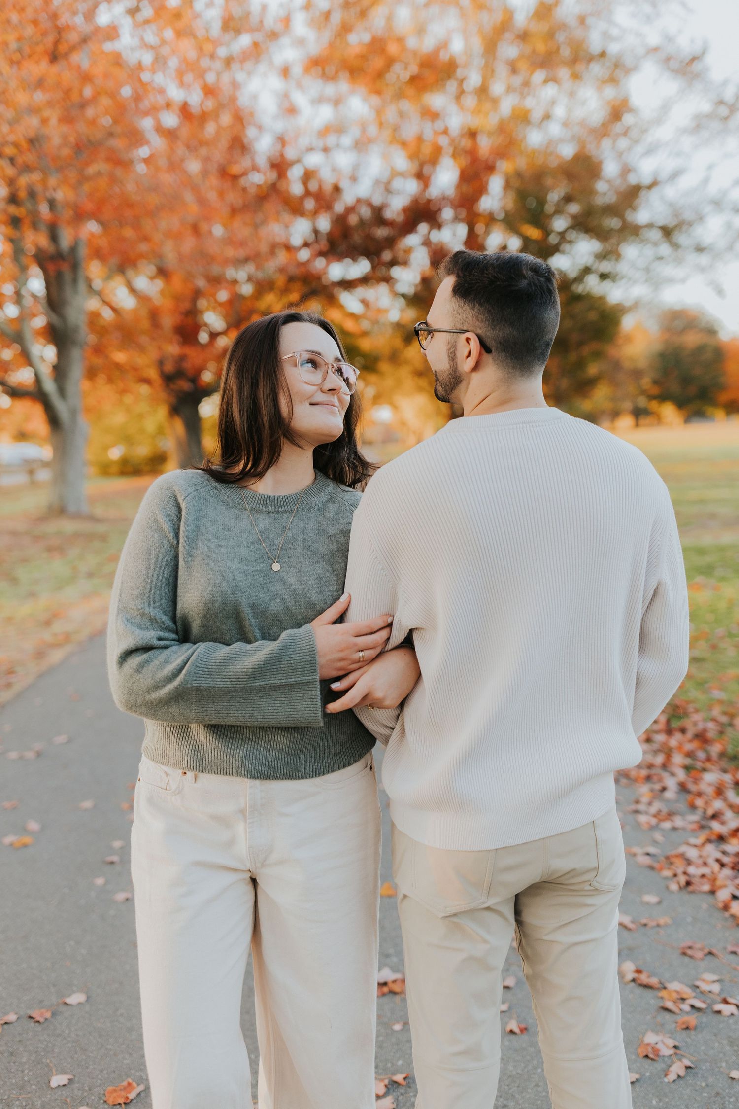 Couple walking hand-in-hand on autumn park path, woman in gray sweater smiling up at man in white sweatshirt.