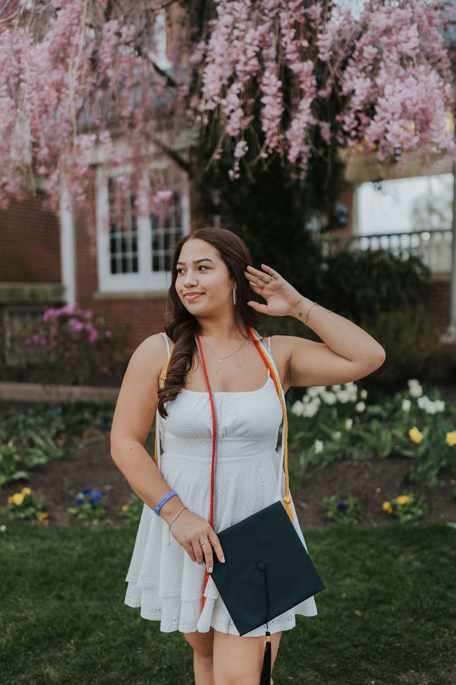Smiling graduate in white dress holds cap under blooming pink cherry blossom tree with colorful honor cords.