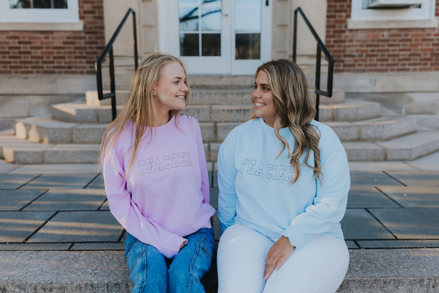Two smiling women in pastel purple and blue sweatshirts sitting together on steps outdoors.