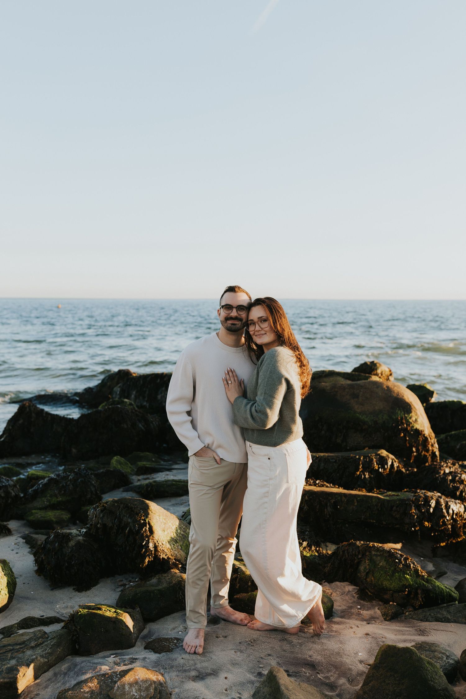 Couple posing on rocky beach at sunset, woman in green sweater leaning against mans chest.