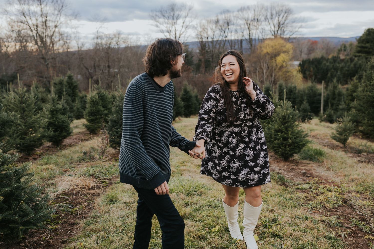 Couple laughs and holds hands while walking through a Christmas tree farm during fall engagement photos.