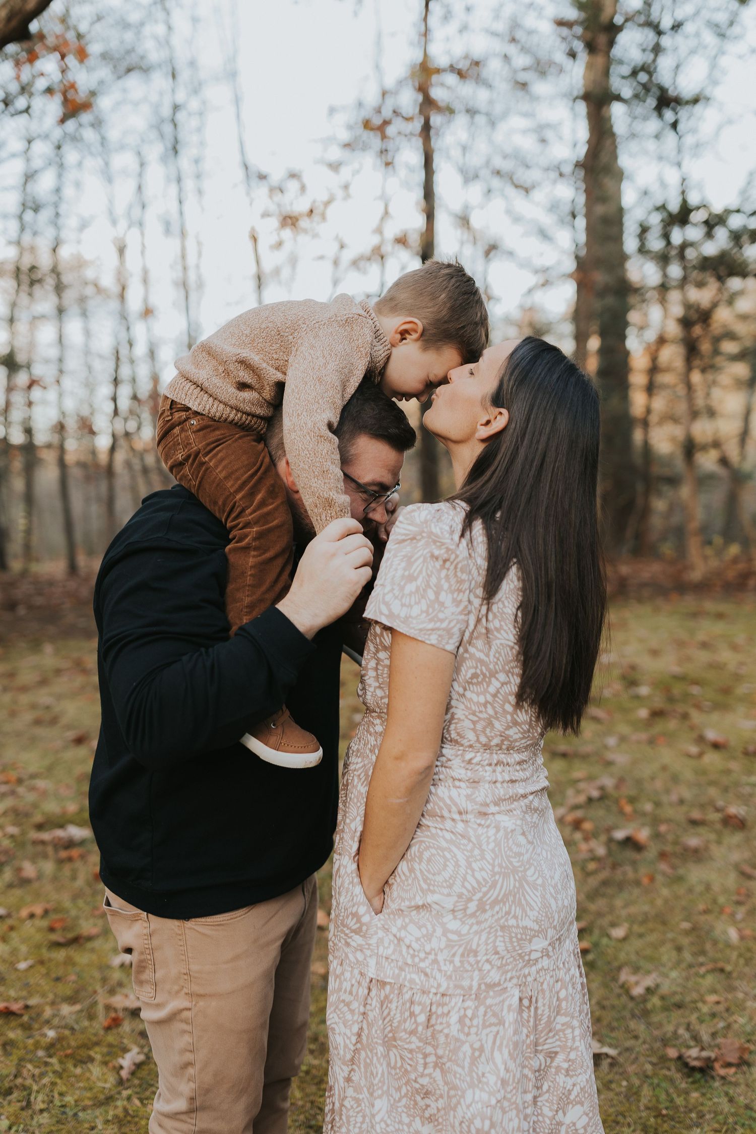 Parents kiss their toddler son in an autumn forest, creating a warm family portrait among fall foliage.