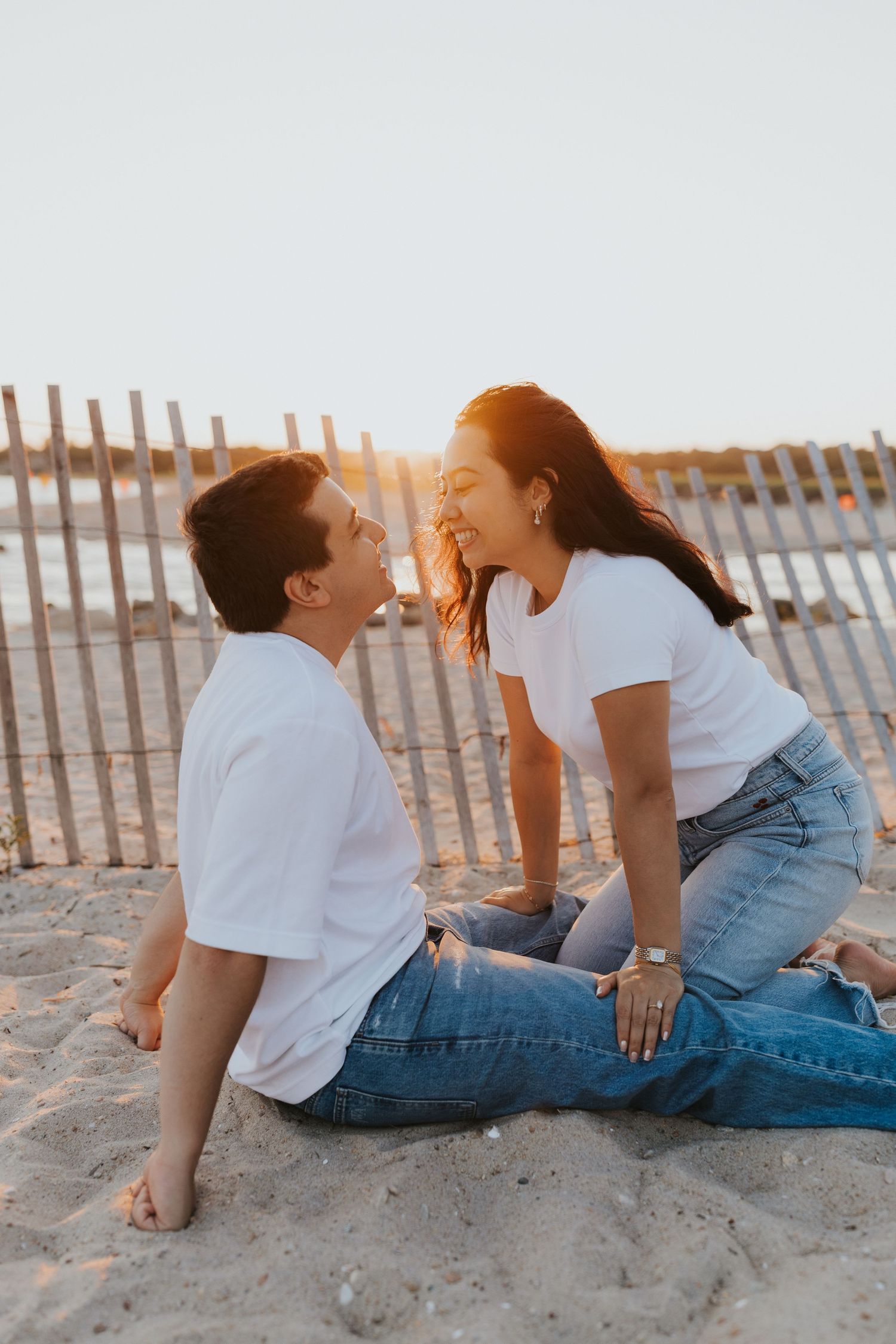 Couple in matching white tees and jeans sitting on sandy beach at golden hour, smiling at each other near a wooden fence.