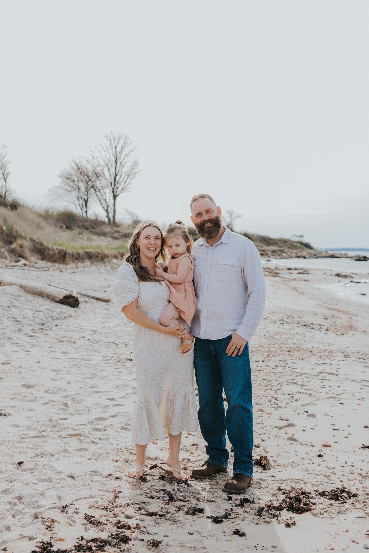 Happy family of three posing on sandy beach, pregnant mother holding toddler while father stands beside them.