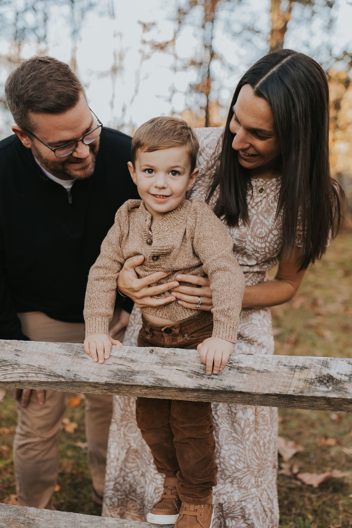 Parents lovingly surround their toddler son as he stands on a rustic wooden fence outdoors.