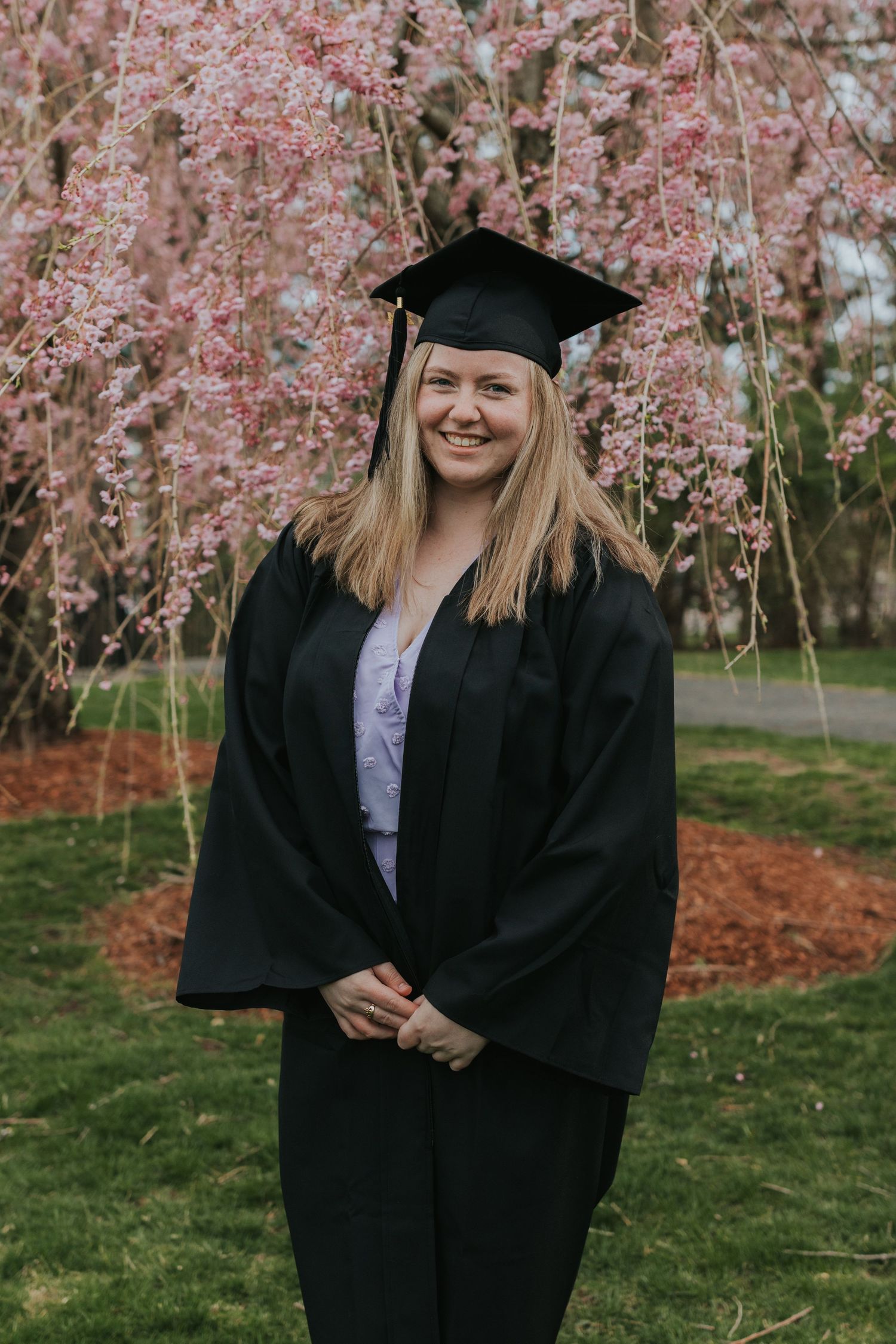 Smiling graduate in black cap and gown poses before blooming pink cherry blossom tree in spring.