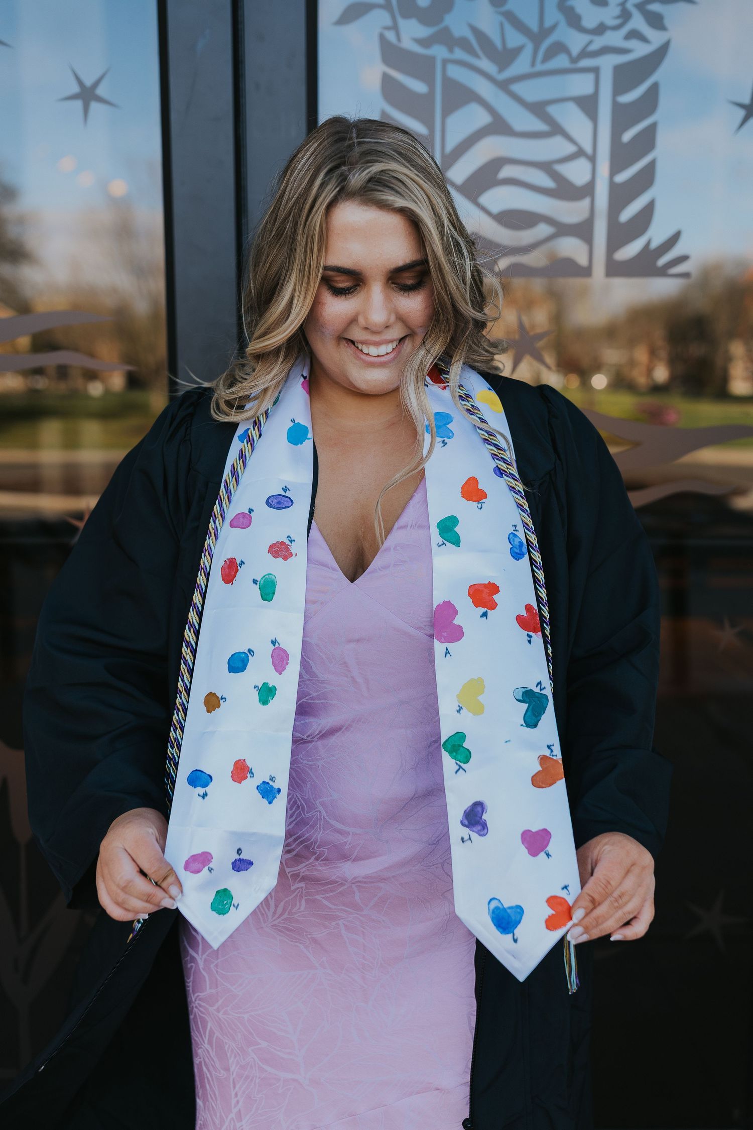 Graduate in black gown smiling at colorful handprint graduation stole.