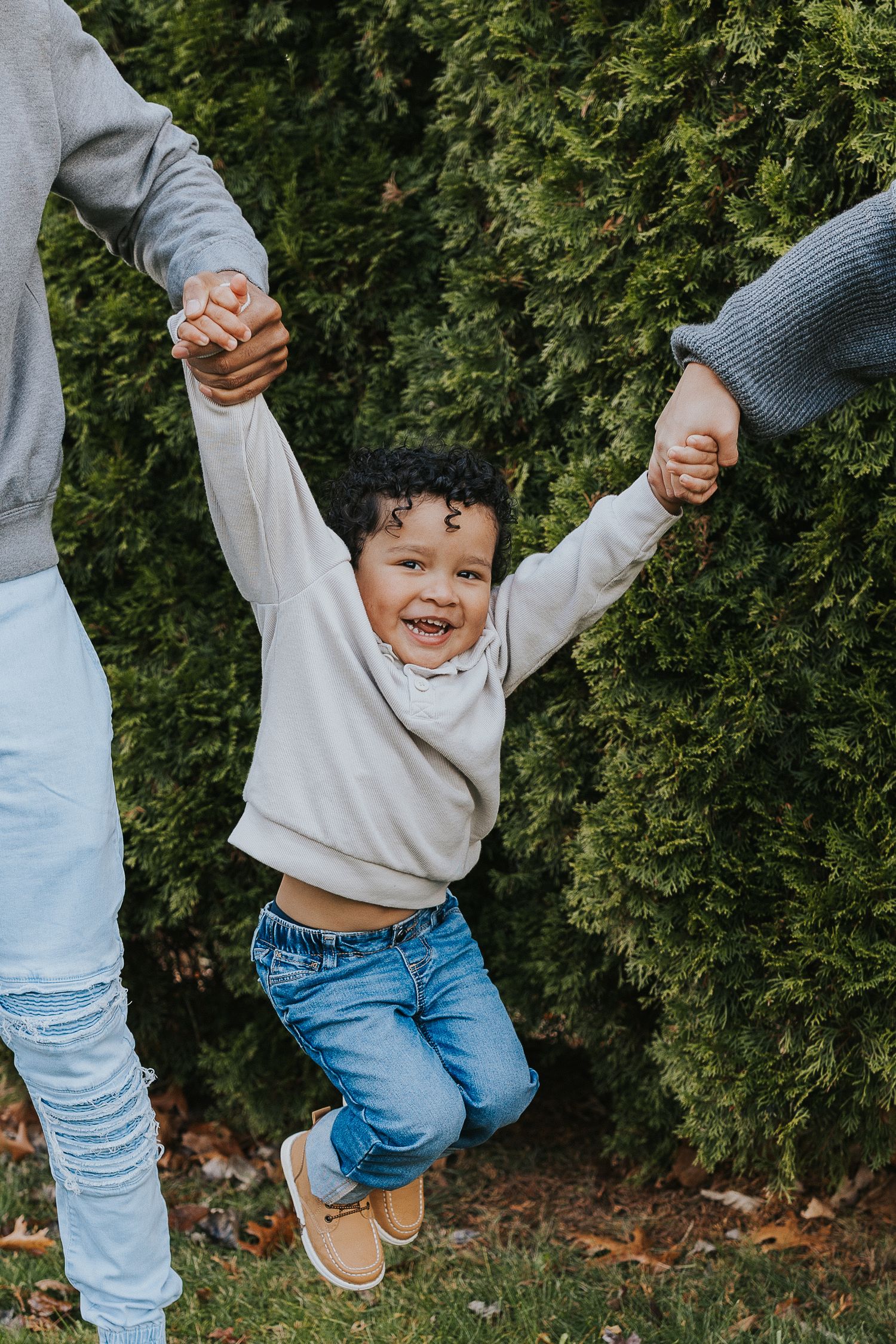 Happy toddler boy being swung by his parents hands in front of green hedges outdoors.