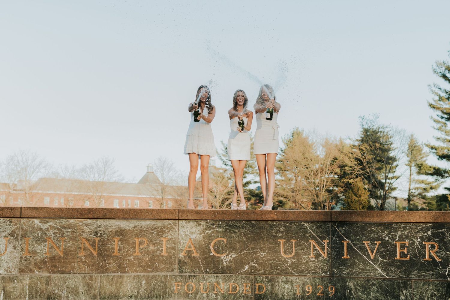 Three female graduates in white dresses stand atop a university sign wall celebrating their graduation outdoors.