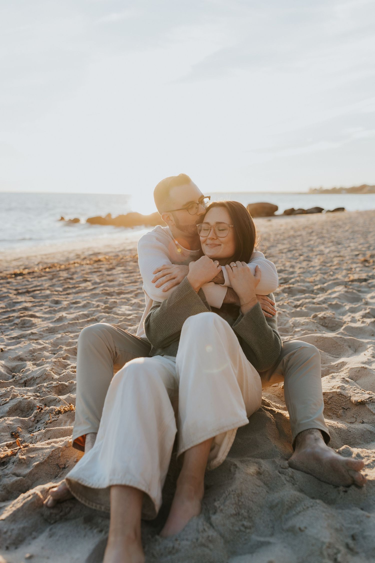 Couple cuddling on sandy beach at golden hour sunset, sitting together peacefully by the ocean.