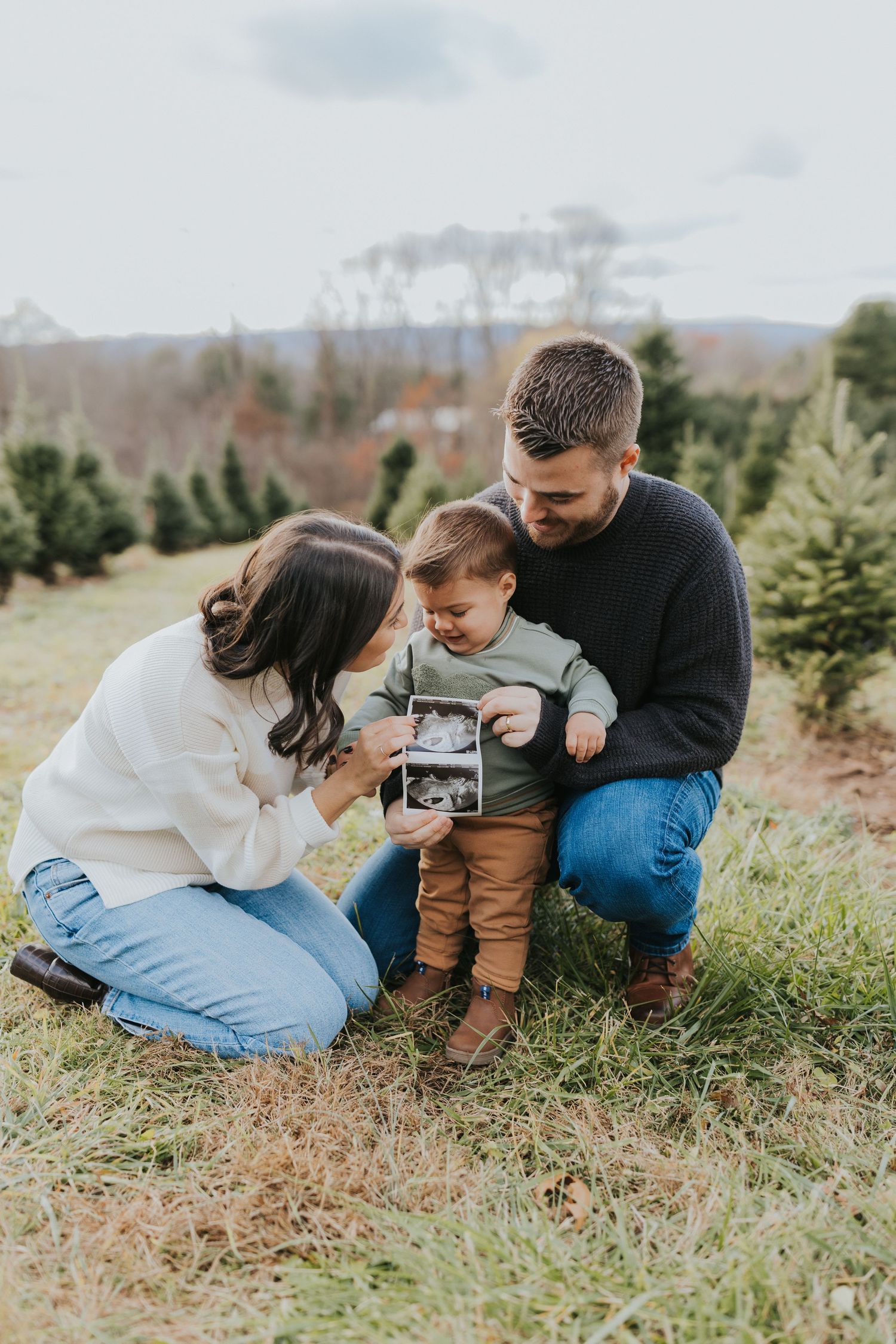 Family of three at Christmas tree farm, toddler holding ultrasound photo for pregnancy announcement.