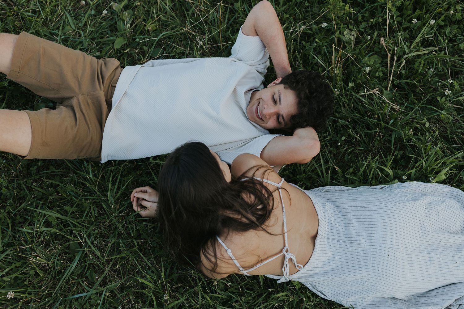 Couple lying in green grass looking at each other, aerial view of relaxed outdoor portrait session.