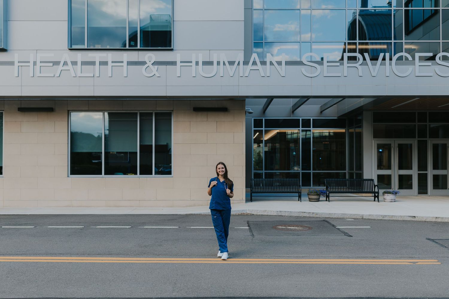 Nursing student in blue scrubs walks outside Health and Human Services building.