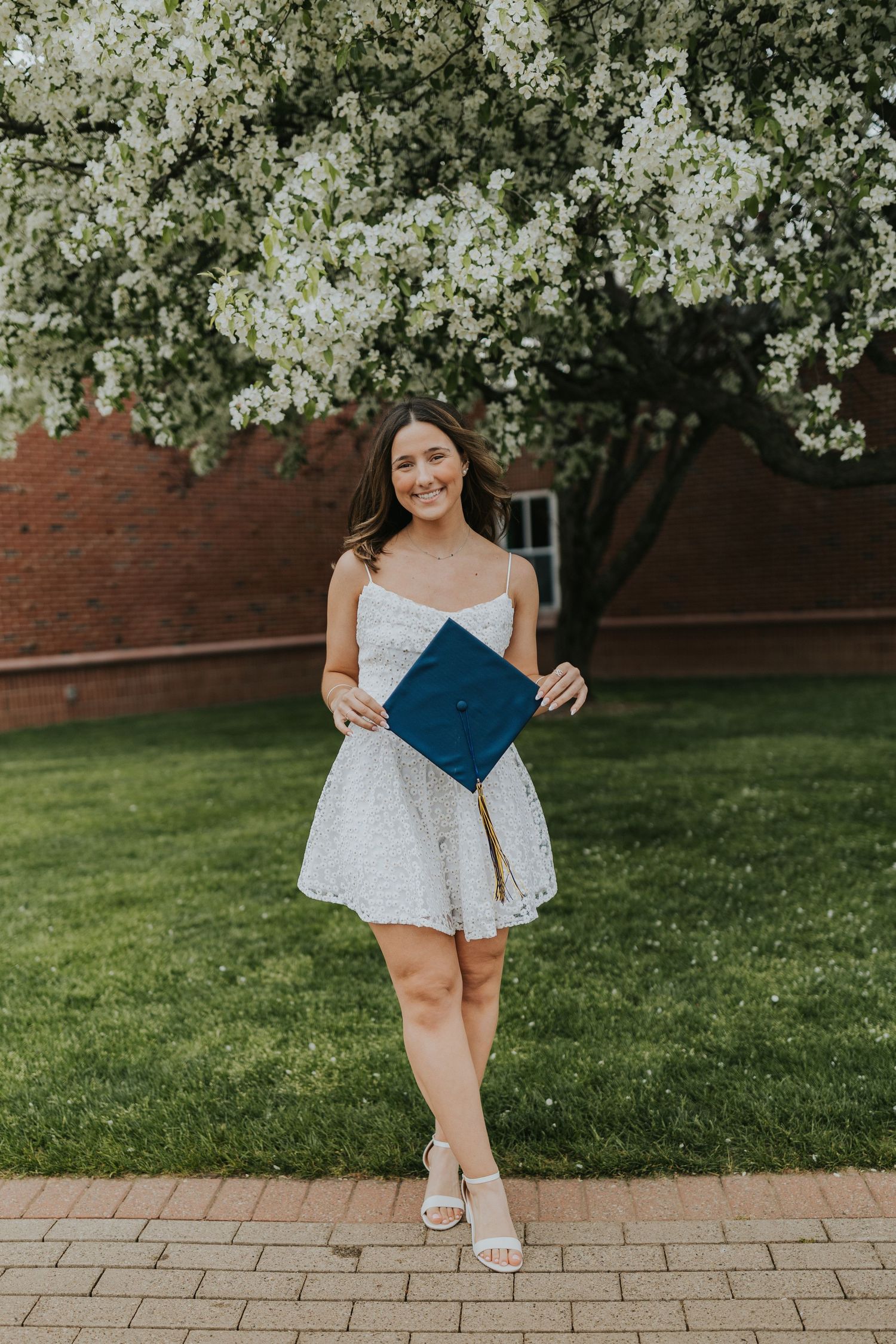 Smiling graduate in white dress holds navy mortarboard cap under flowering white tree on campus.