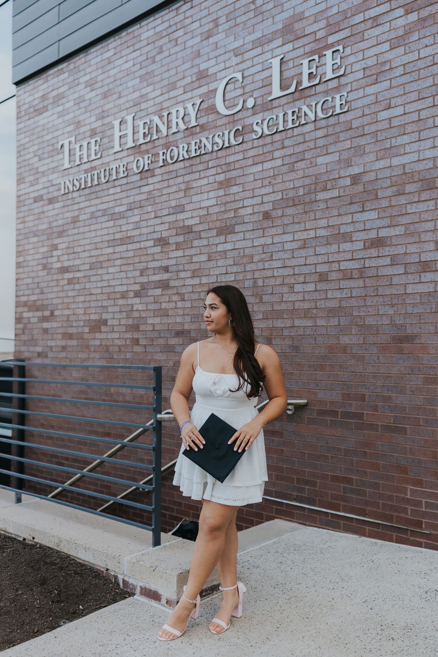 Graduate in white dress poses beside Henry C. Lee Institute of Forensic Science brick wall.