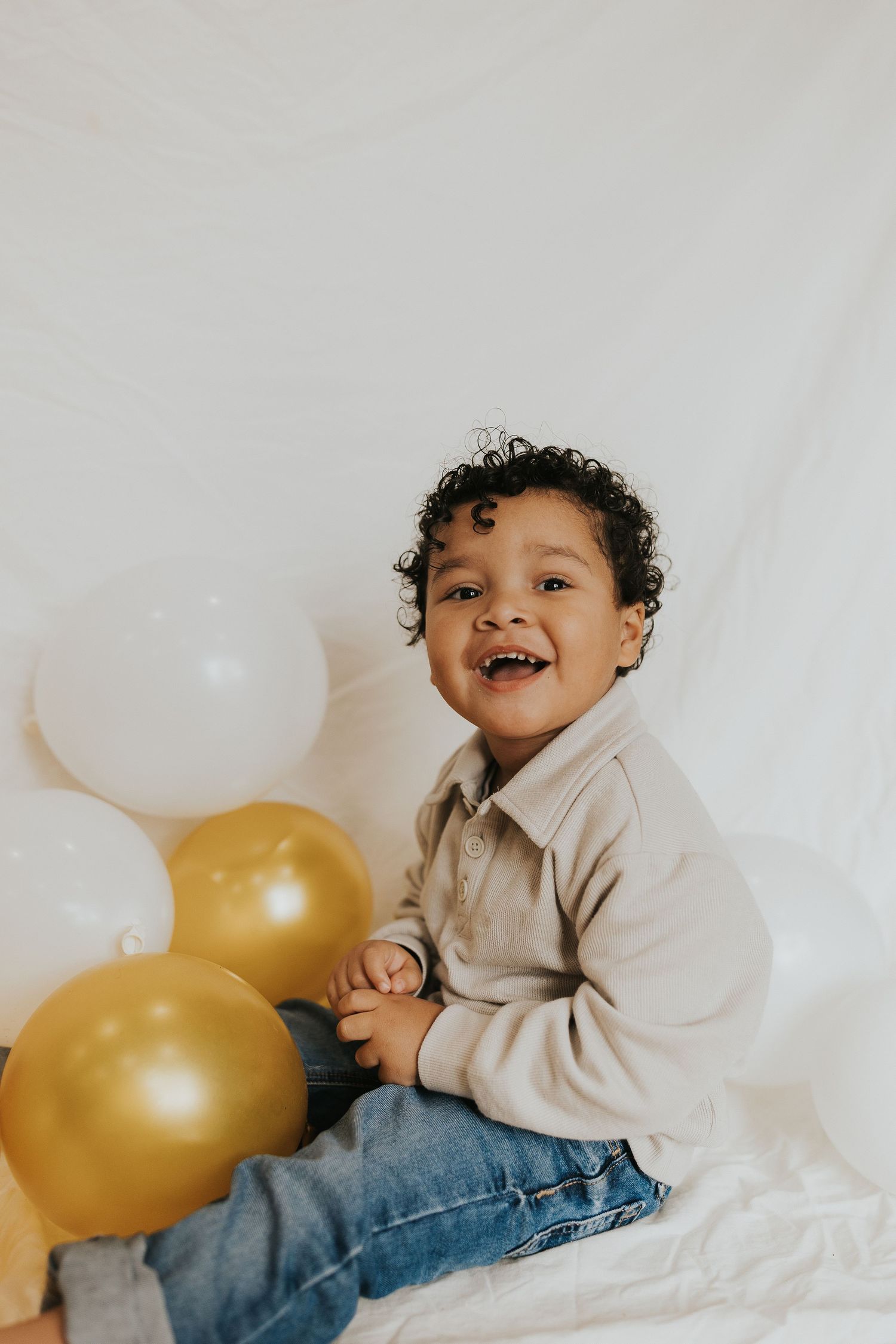 A smiling toddler with curly hair sits happily among gold and white balloons against white backdrop.