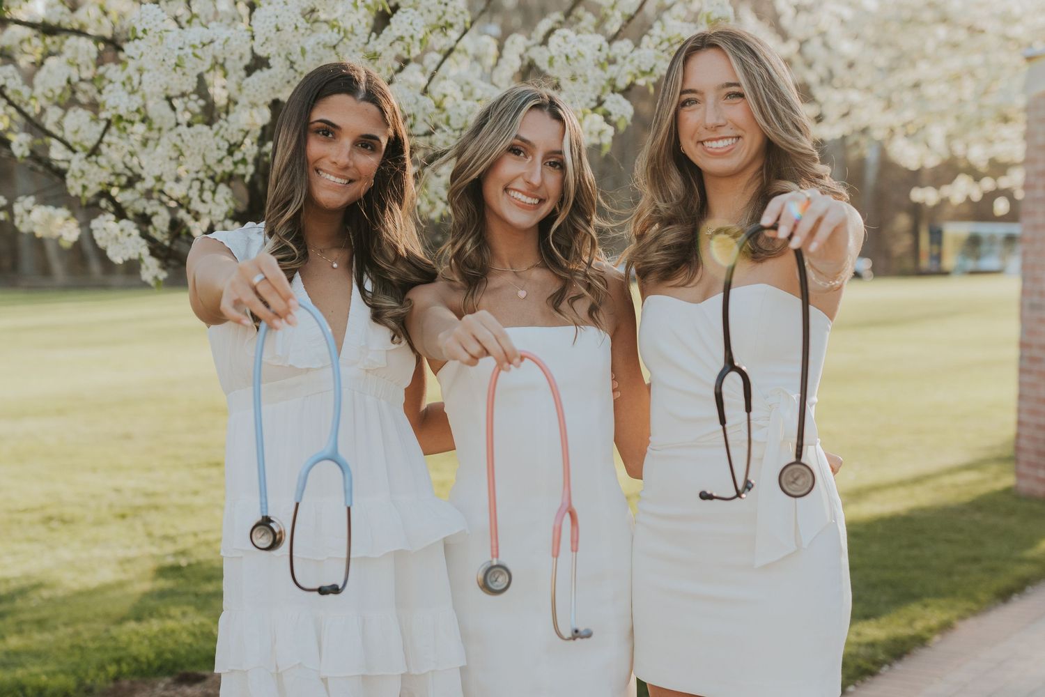 Three nursing graduates in white dresses proudly displaying their colorful stethoscopes near blooming trees.