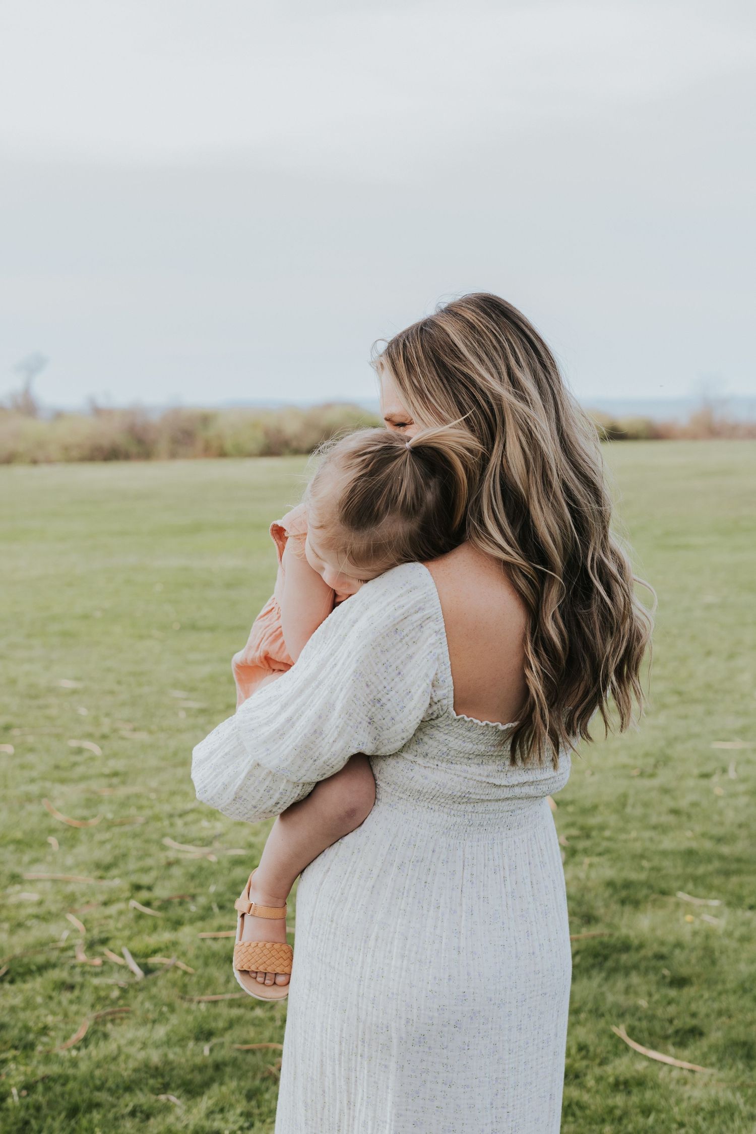 Mother with long wavy hair cradles sleeping toddler in orange dress in open green field.