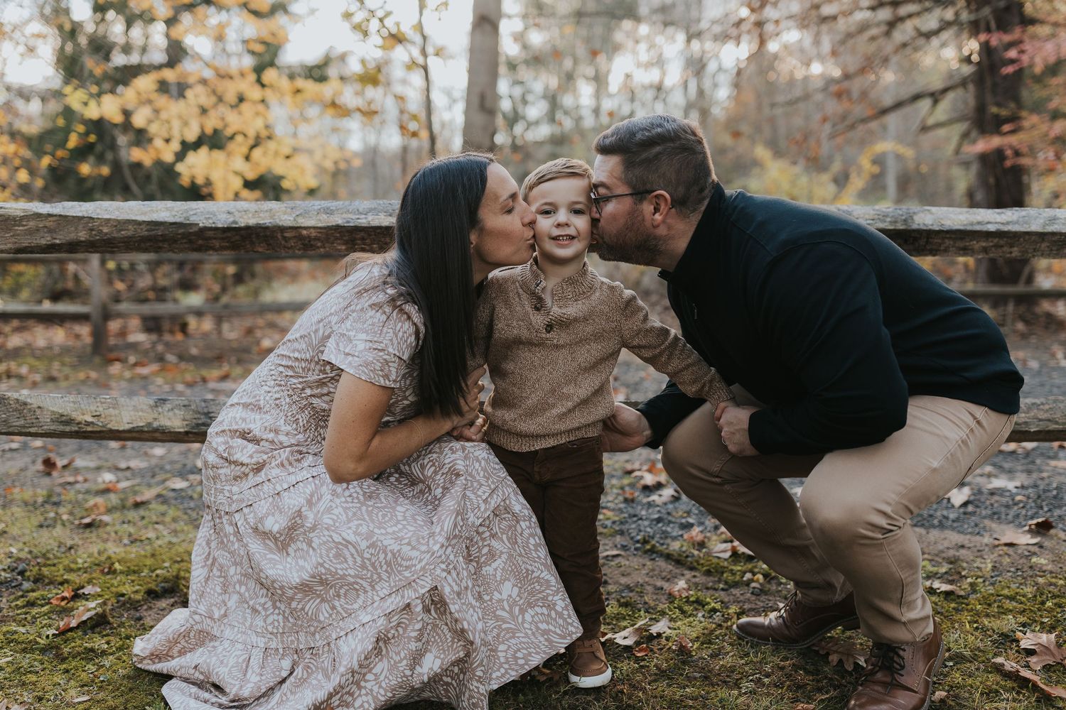 Two parents lovingly kiss their smiling toddler son on the cheeks outdoors in fall foliage.