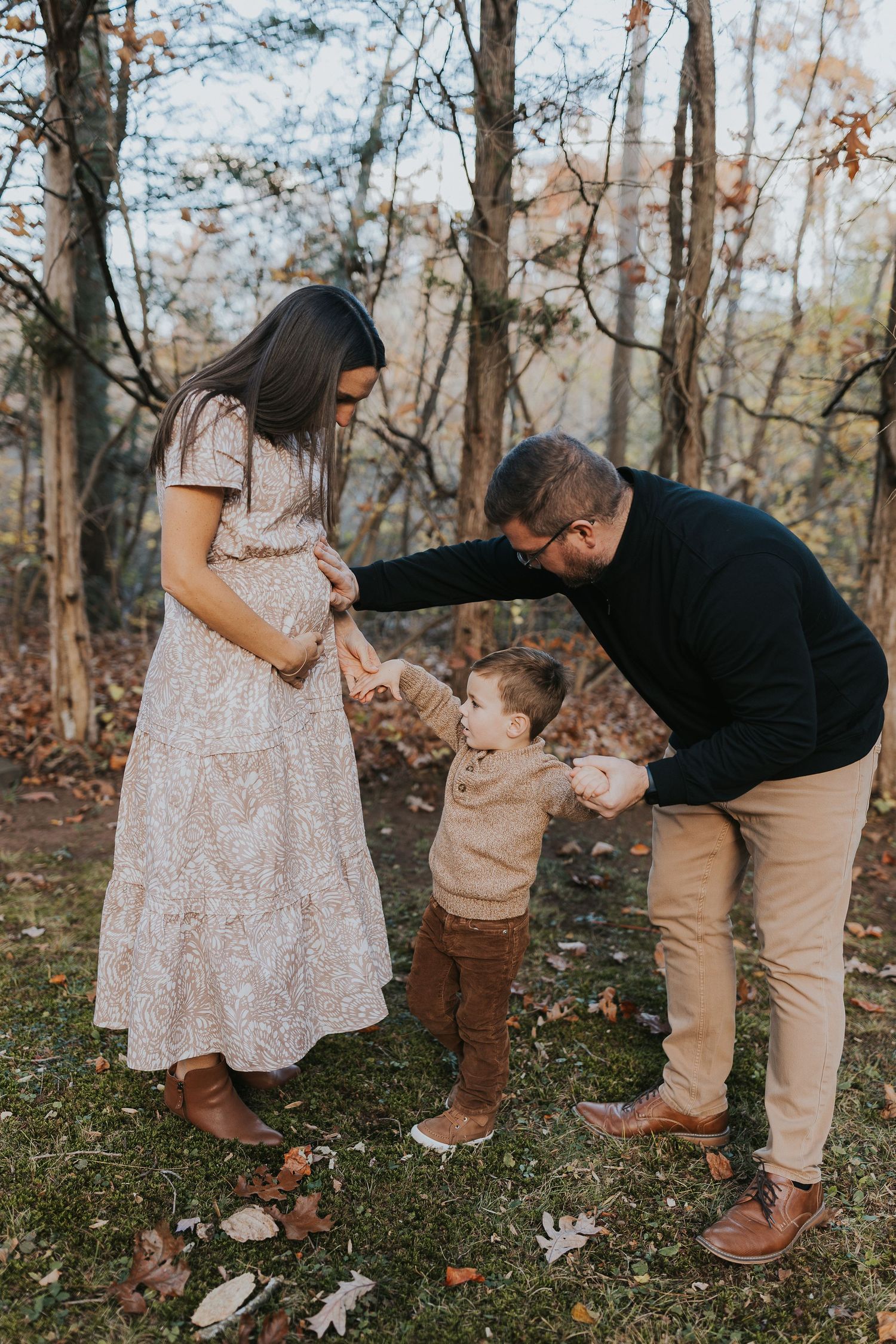 Expecting parents hold hands with their toddler son in a colorful autumn forest setting.