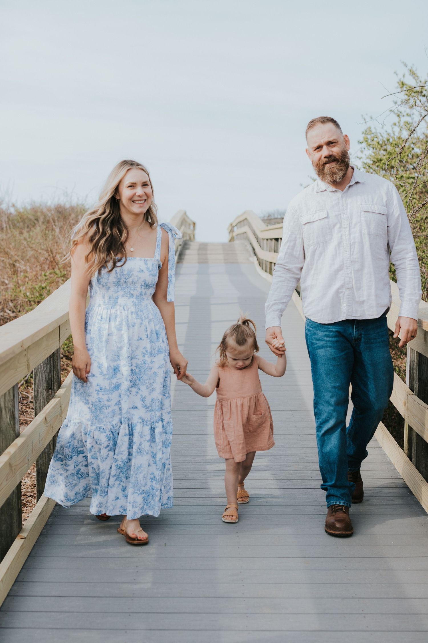 Joyful family of three walks on wooden boardwalk holding hands, daughter in pink dress.