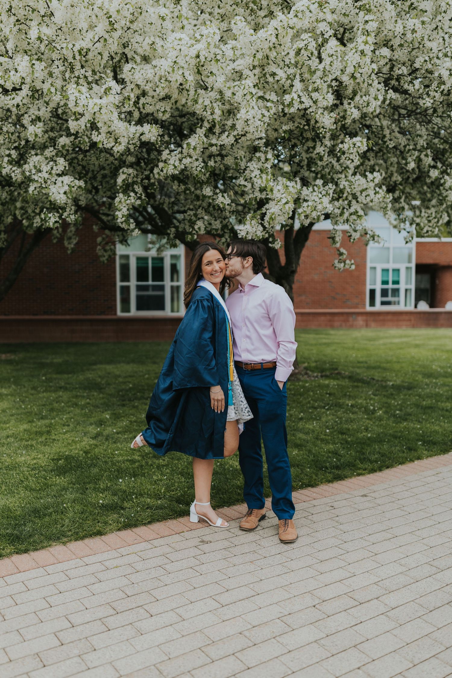 Graduate in navy gown shares a romantic moment with partner under blooming white spring tree.