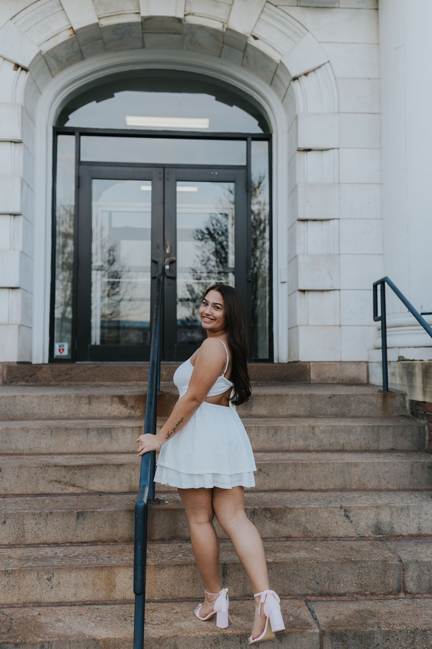 Smiling young woman in white mini dress poses on stone steps of classic building.