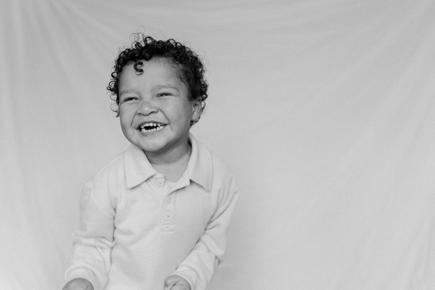 A laughing young boy with curly hair poses for a cheerful black and white portrait photo.
