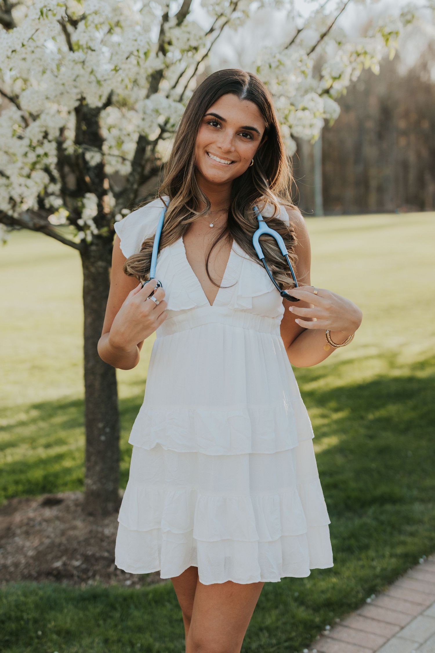 Smiling nursing student in white tiered dress holding stethoscope near blooming cherry blossom tree.