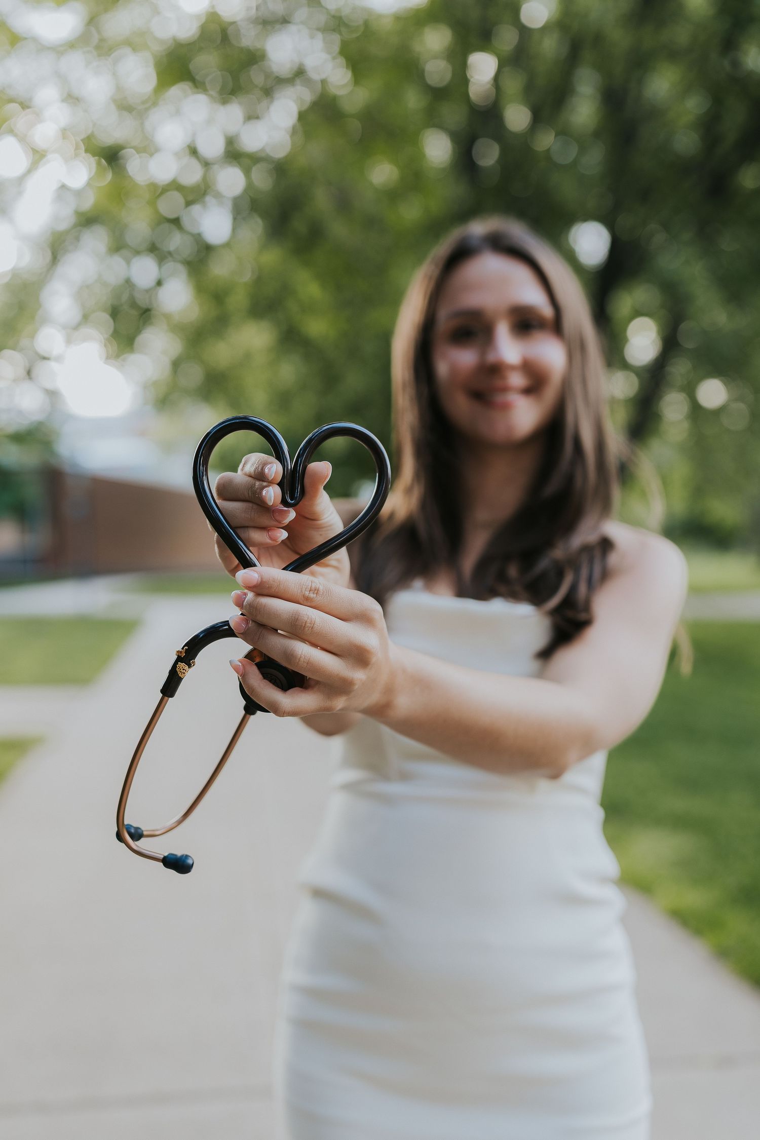 Nursing graduate holds stethoscope shaped into heart toward camera outdoors.