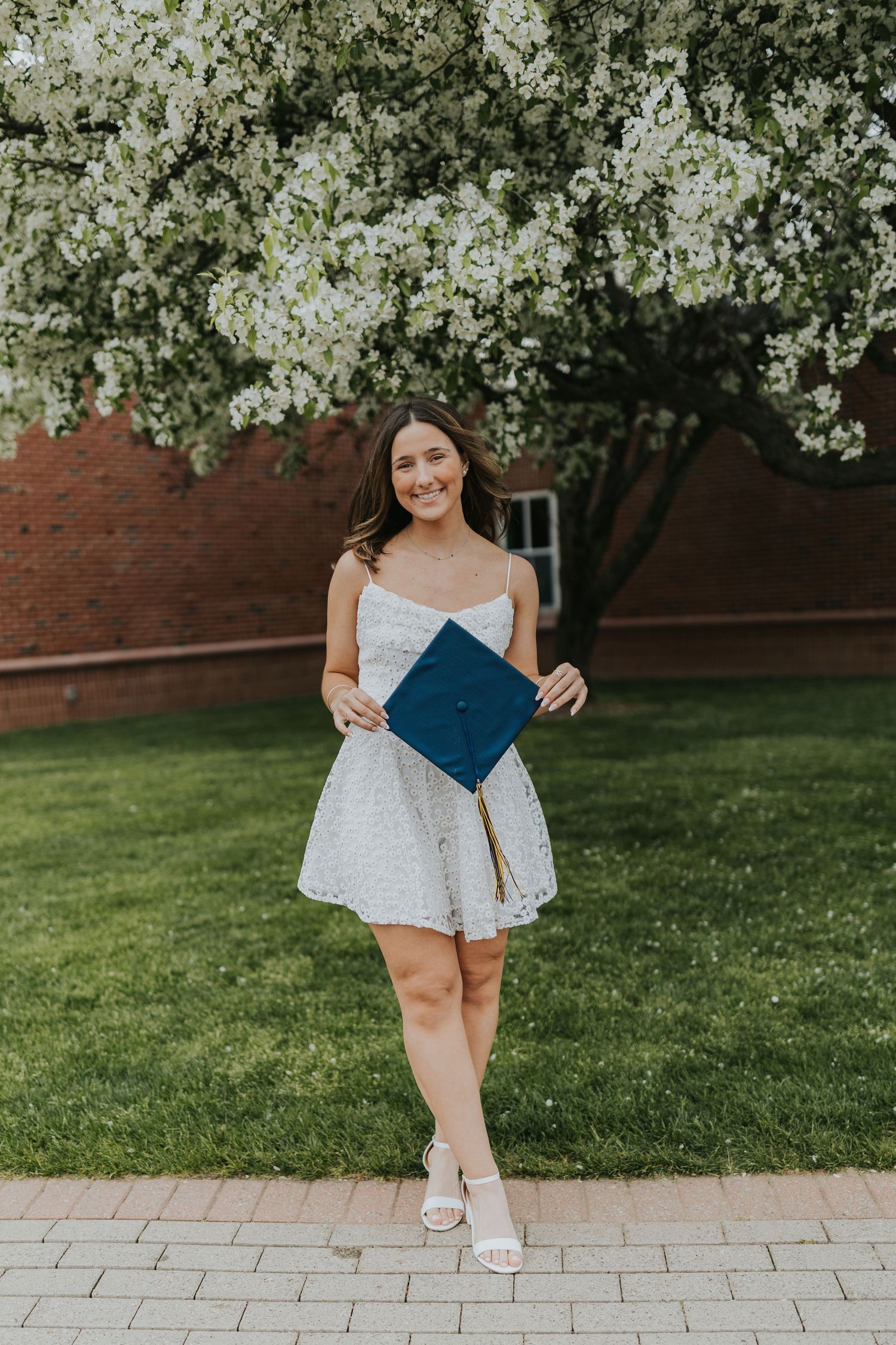 Smiling graduate in white dress holding navy mortarboard under blooming white flowering tree.