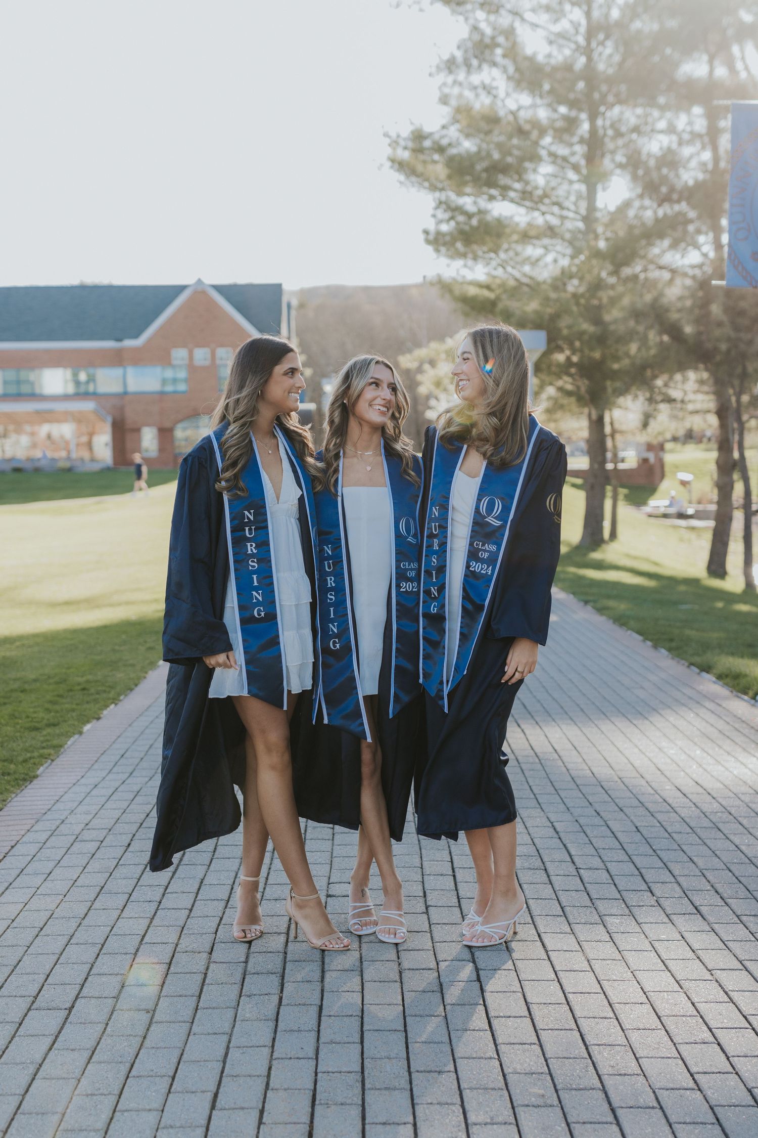 Three nursing graduates in navy gowns and stoles walk together on sunlit campus brick pathway.