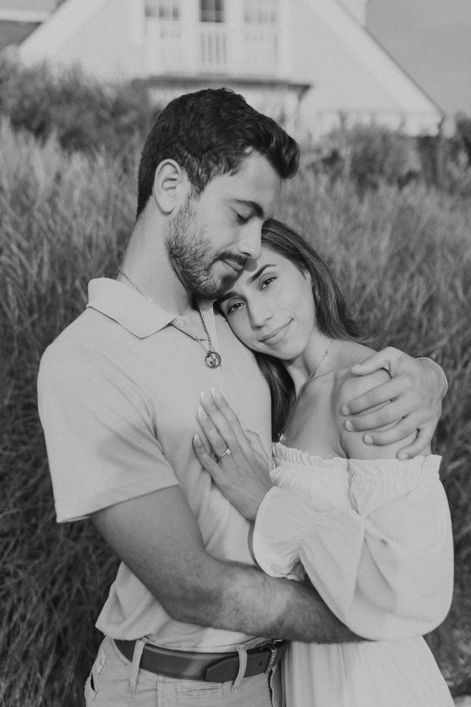 Black and white photo of couple sharing a tender embrace in a field, woman wearing engagement ring.