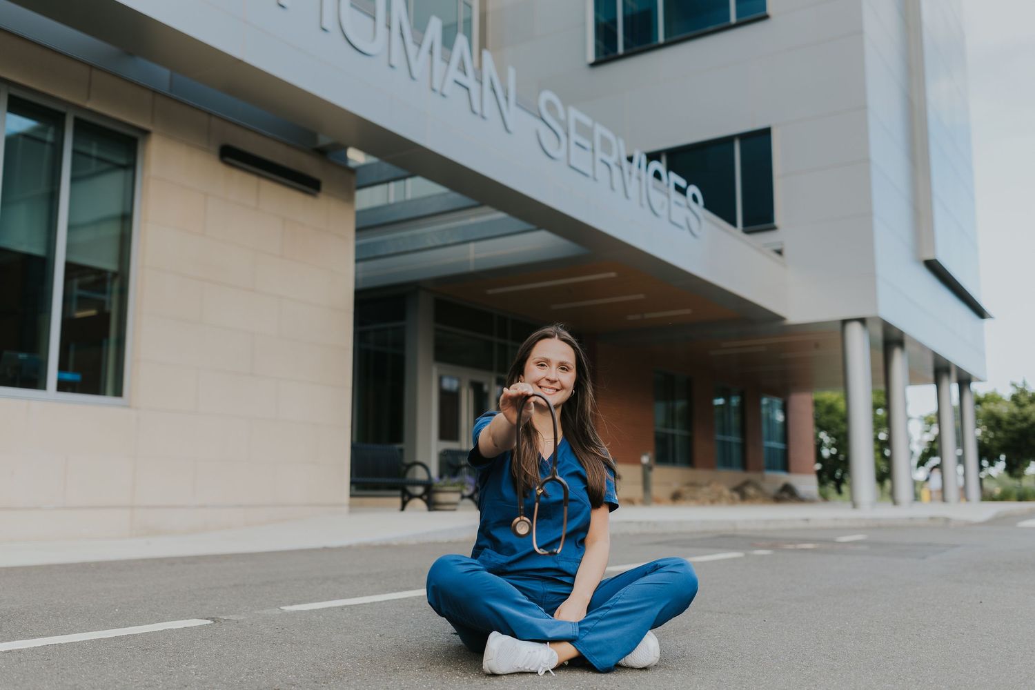 Smiling nurse in blue scrubs sitting cross-legged outside a Human Services building, talking on phone.