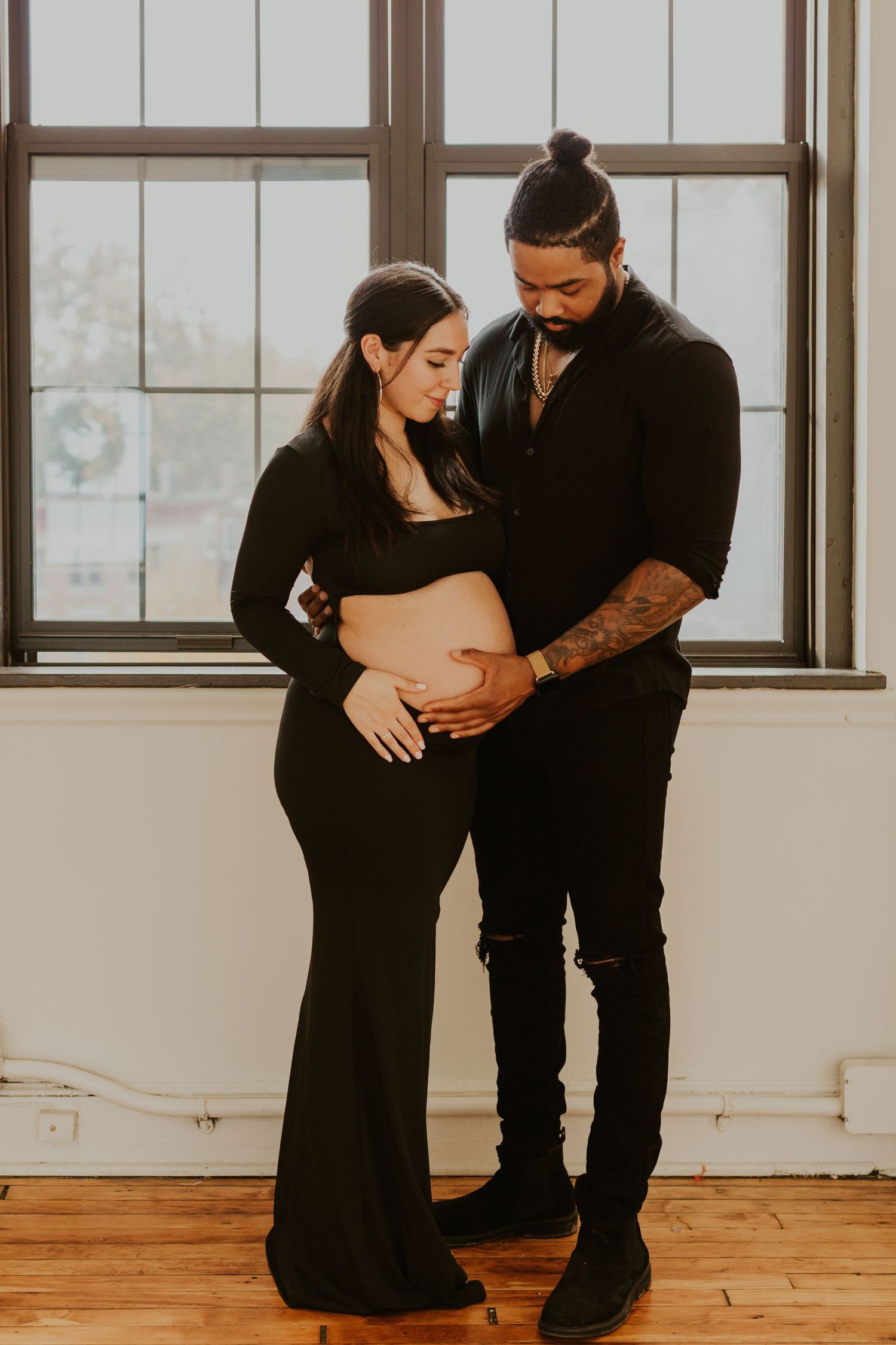 Couple in matching black outfits tenderly looking at pregnant belly during indoor maternity photoshoot.